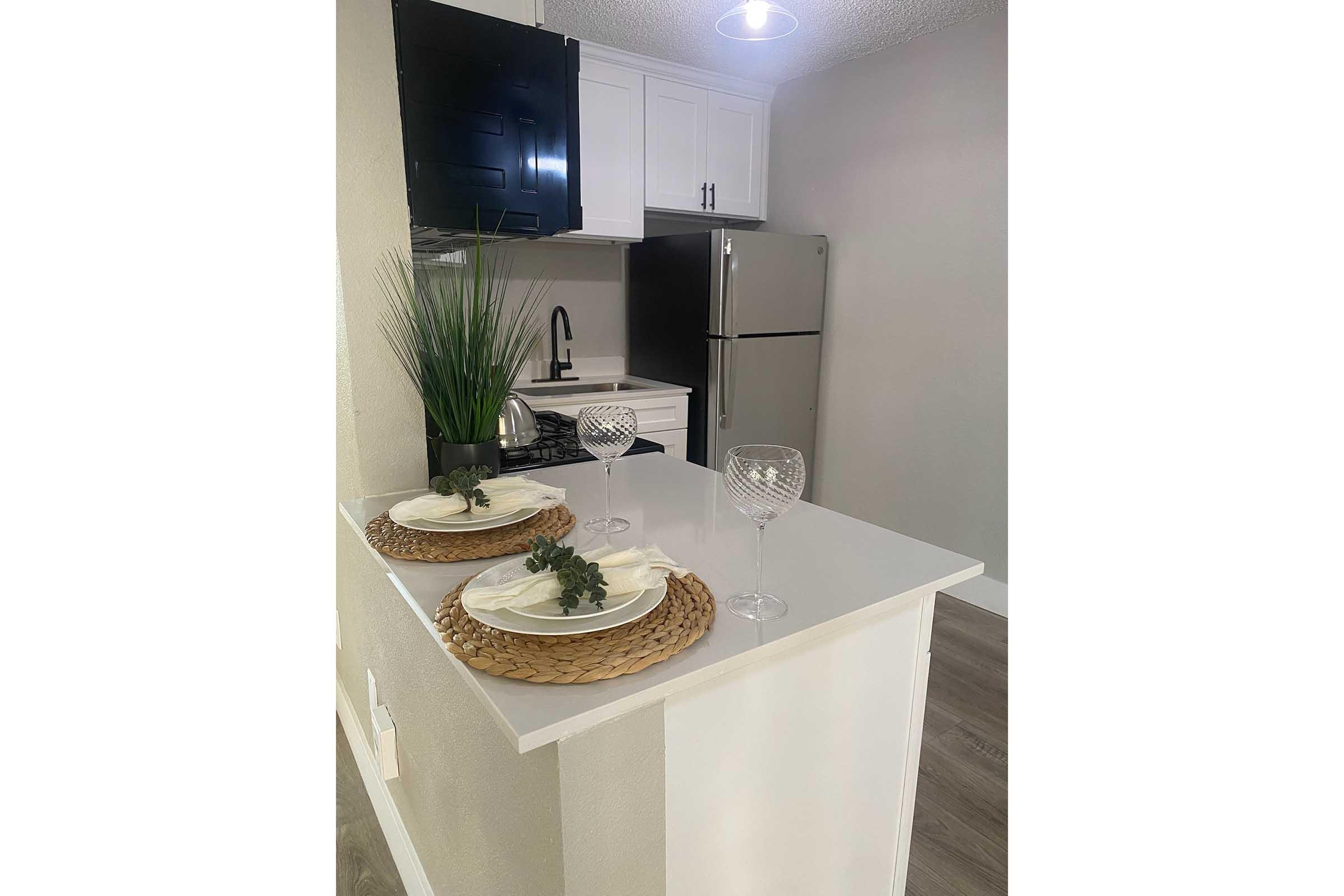 A modern kitchen featuring a white countertop with a decorative setting that includes two wine glasses, plates, and greenery. The background shows sleek cabinets, stainless steel appliances, and a stylish faucet, creating a contemporary and inviting atmosphere.