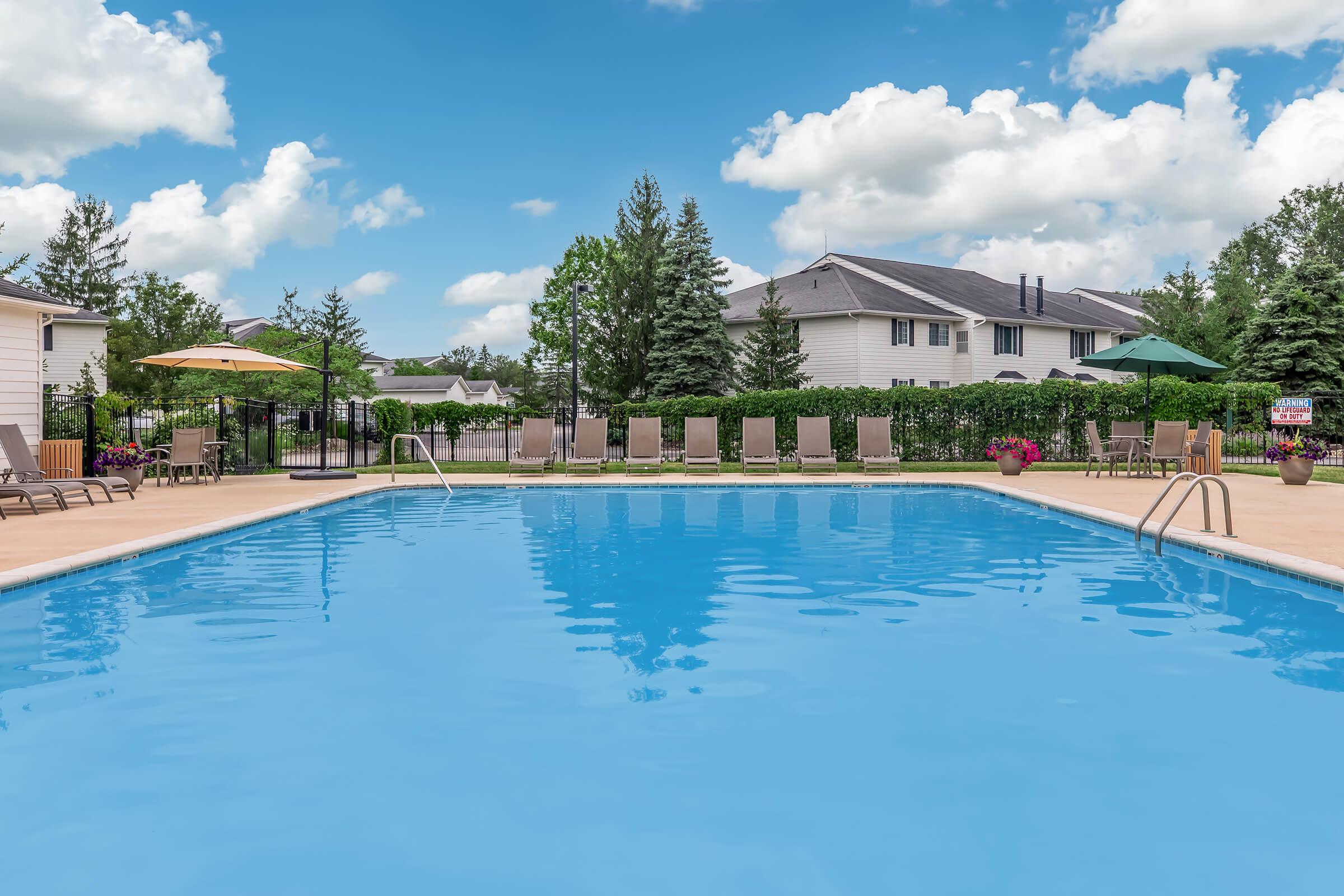 A clear blue swimming pool surrounded by lounge chairs and umbrellas, set against a backdrop of green trees and a residential area with houses under a partly cloudy sky. The scene conveys a relaxing outdoor atmosphere.