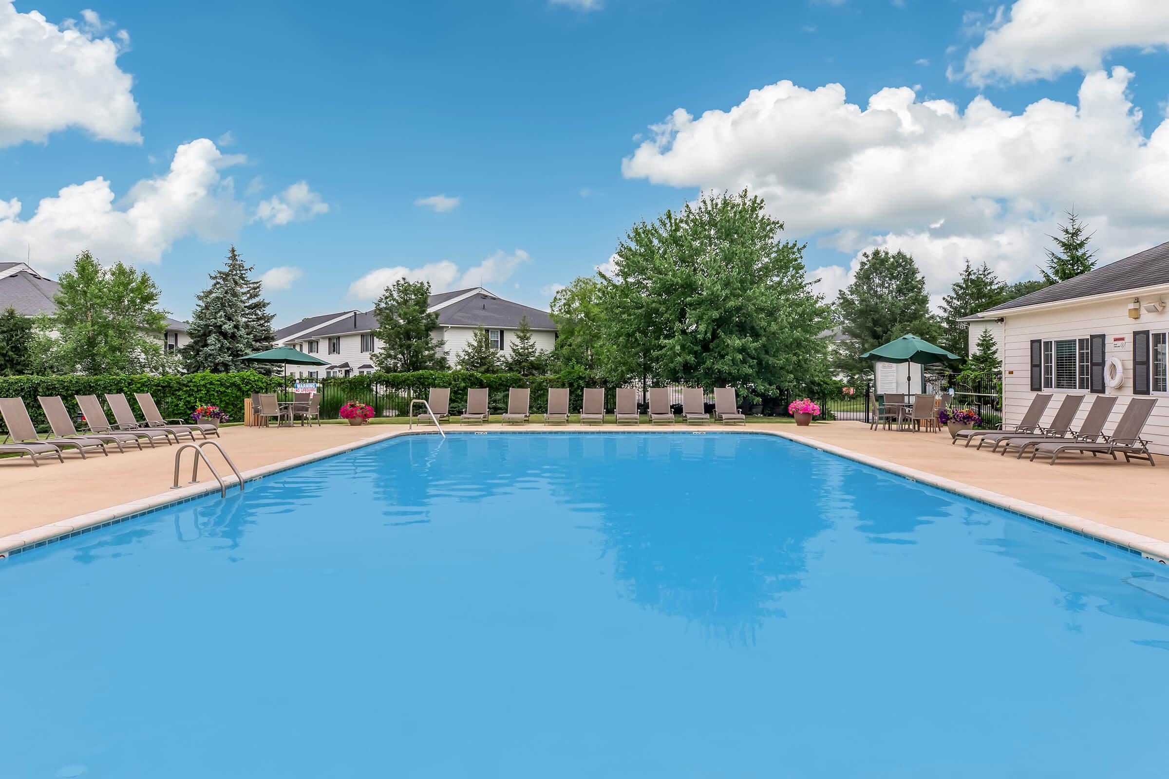 A bright blue swimming pool surrounded by lounge chairs, with green trees and shrubs in the background. The sky is partly cloudy, creating a serene atmosphere. Some umbrellas provide shade near the pool area, and flower pots add a touch of color to the scene.