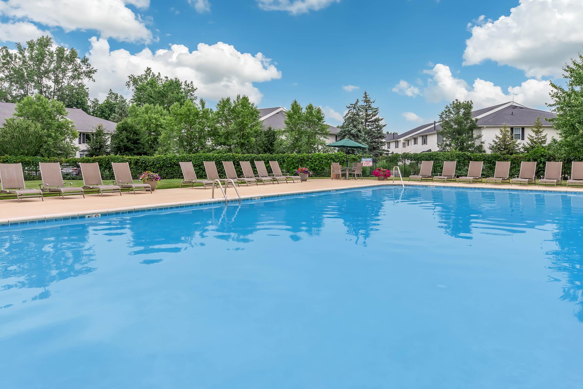 A large, clear swimming pool surrounded by lounge chairs under a bright blue sky with fluffy white clouds. Lush greenery and residential buildings are visible in the background, creating a serene and inviting atmosphere.
