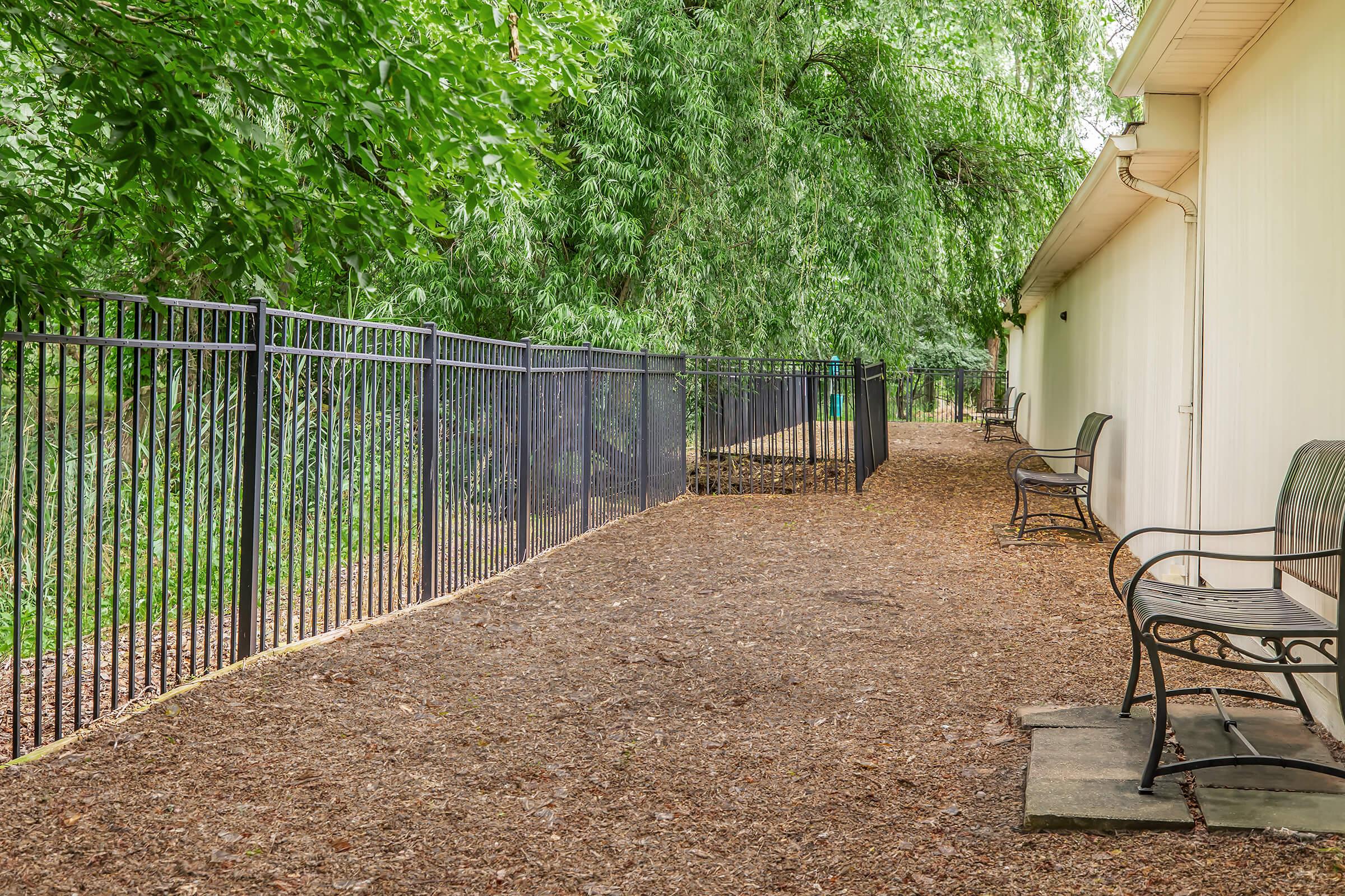 A narrow pathway lined with a black metal fence and benches on either side, surrounded by lush greenery and trees. The ground is covered with brown mulch, creating a natural and serene atmosphere.