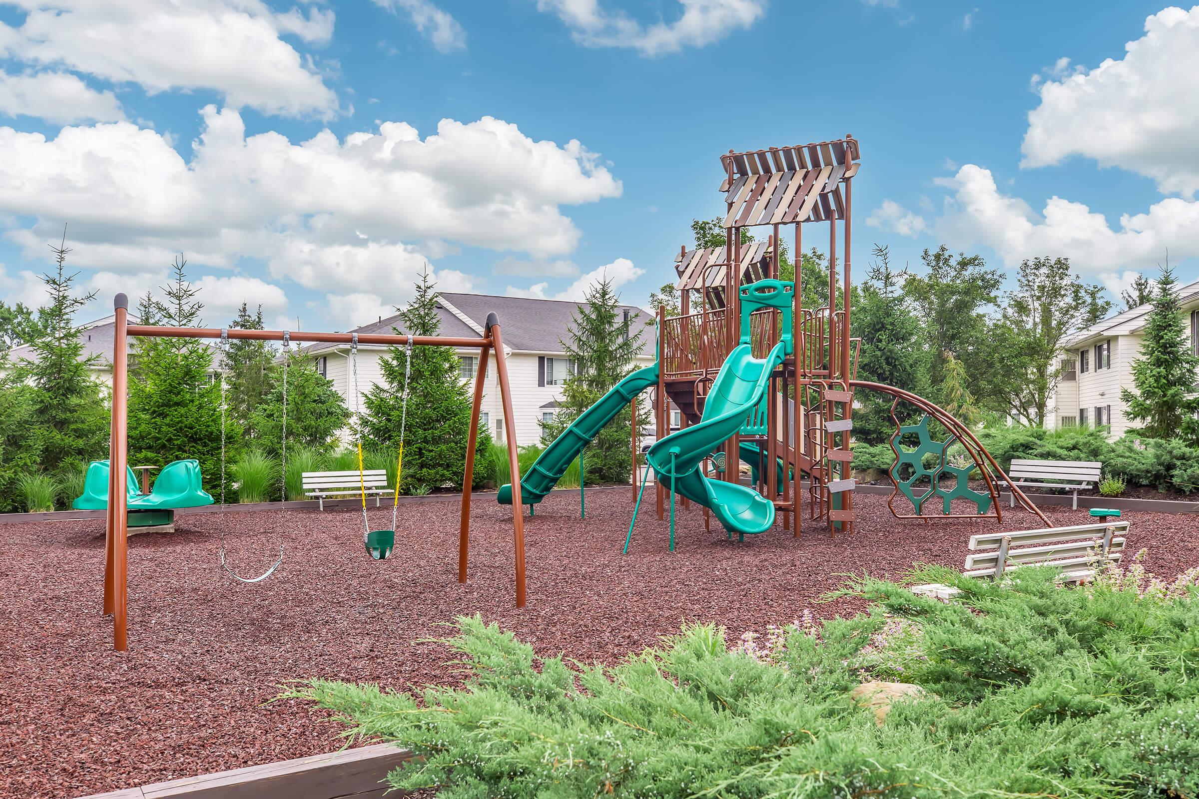 A vibrant playground featuring green slides, swings, and climbing structures, surrounded by mulch and greenery. In the background, residential buildings are visible under a blue sky with fluffy clouds, providing a cheerful and inviting atmosphere for children and families.