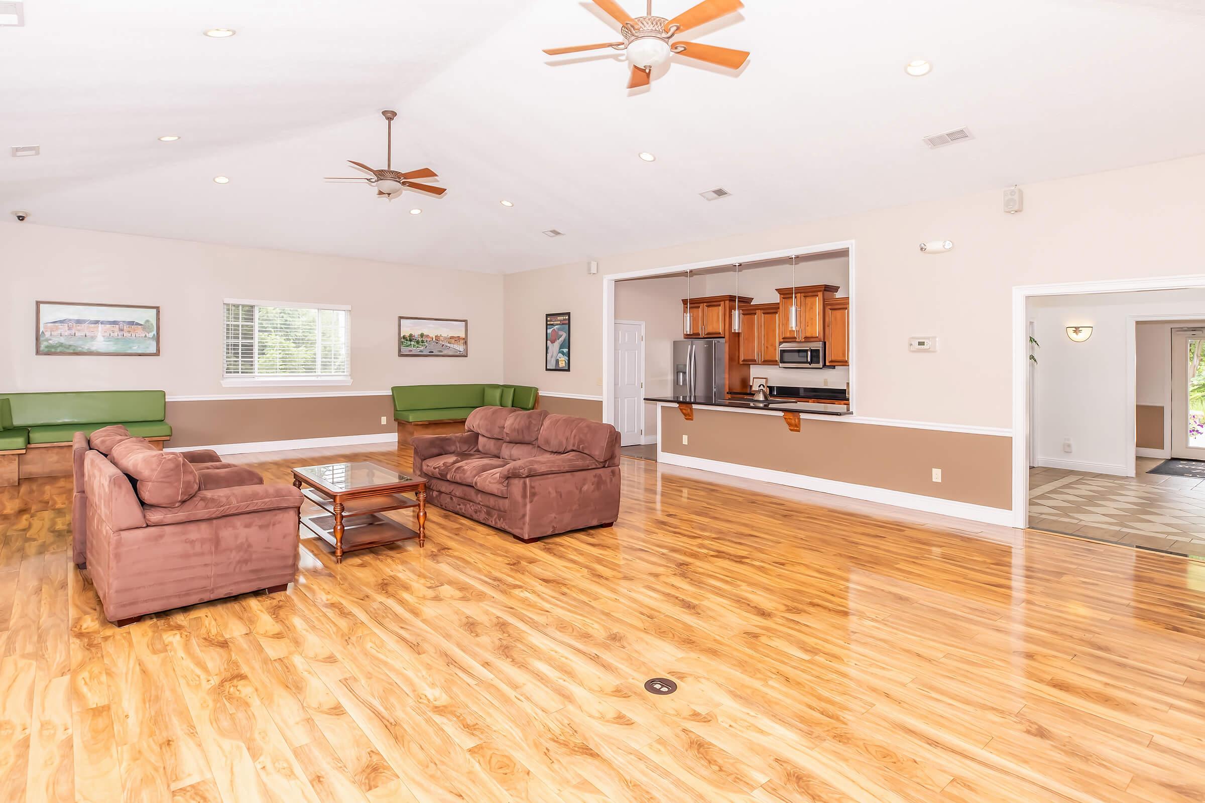 A spacious, well-lit living area featuring two brown sofas and a wooden coffee table. The room has hardwood flooring and is decorated with wall art. There are two ceiling fans and large windows allowing natural light, with green seating along the walls. An open kitchen area is visible in the background.