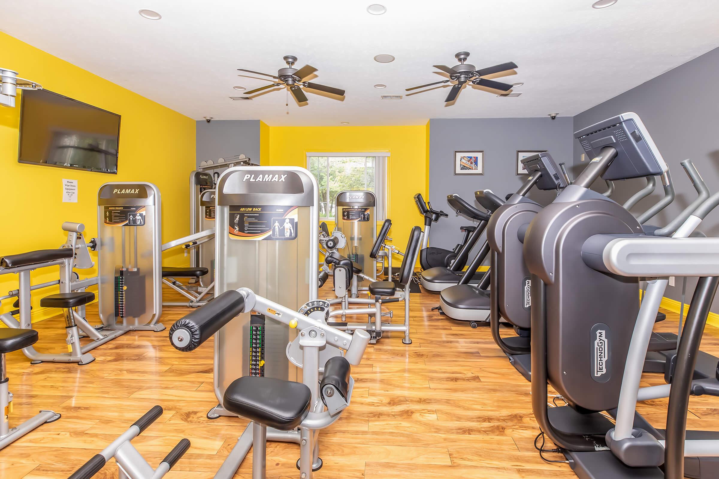 A well-equipped gym with various exercise machines, including weight training equipment, cardio machines, and benches. The room features yellow and gray walls, ceiling fans, and a television, creating a bright and inviting atmosphere for workouts.