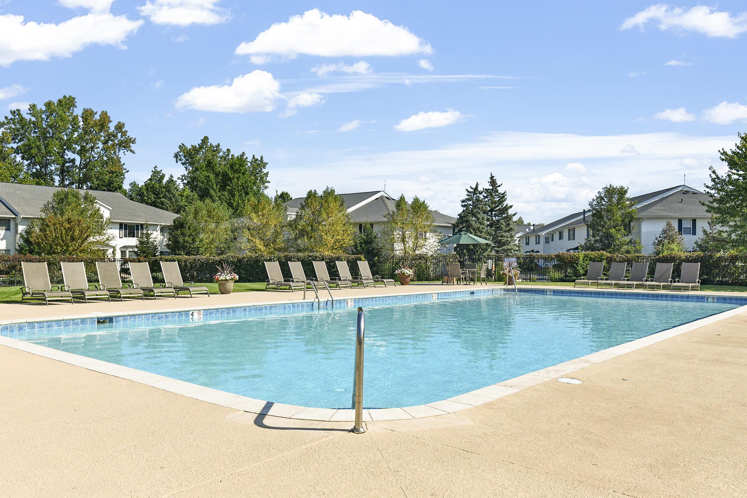 A clear blue swimming pool surrounded by lounge chairs and well-maintained greenery. There are several trees and residential buildings in the background under a bright, sunny sky with a few clouds. The pool area appears inviting and spacious, perfect for relaxation and leisure activities.