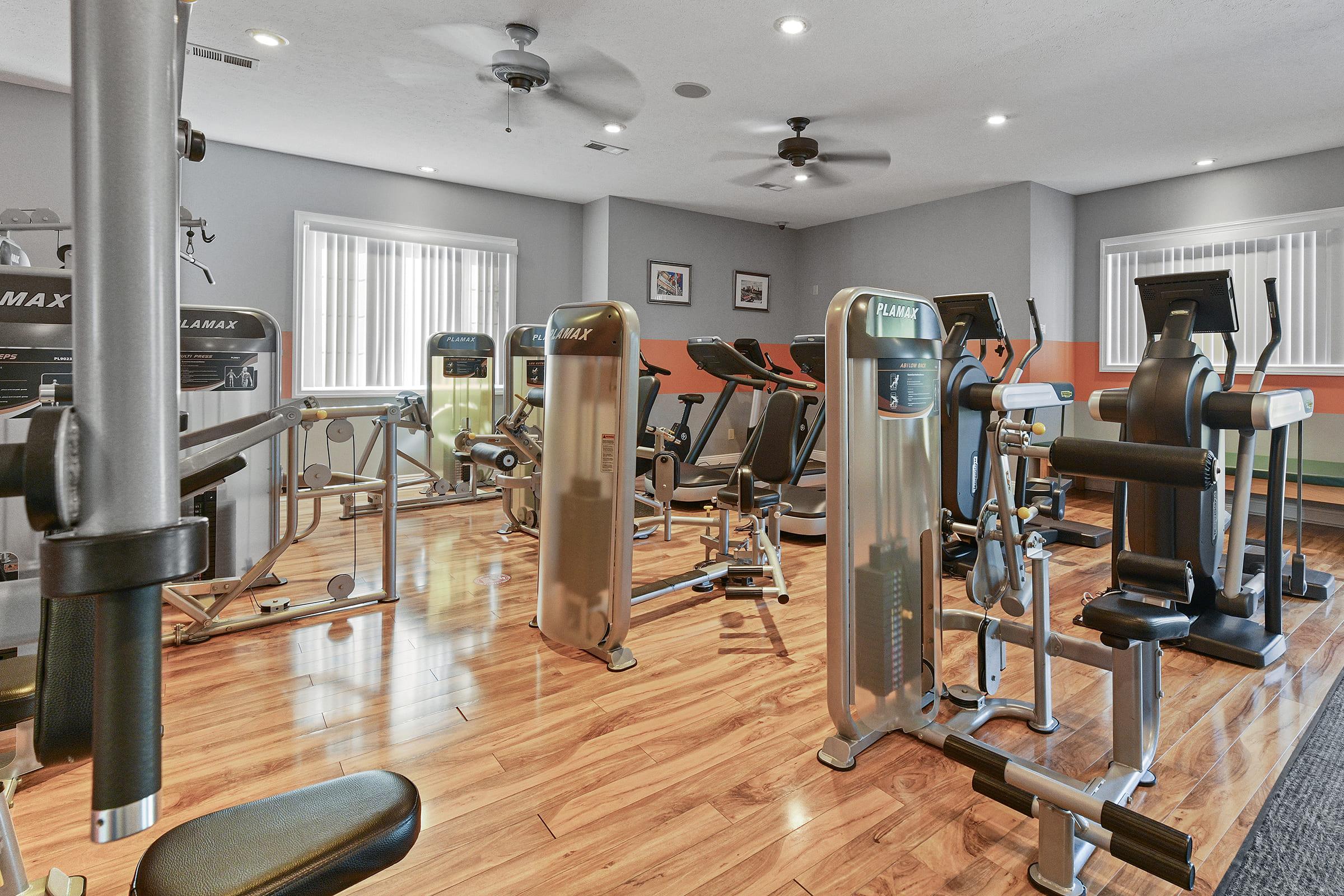 A modern gym interior featuring various exercise machines, including cardio equipment and weight machines. The space has wooden flooring, bright lighting, and large windows allowing natural light, creating a welcoming atmosphere for workouts.