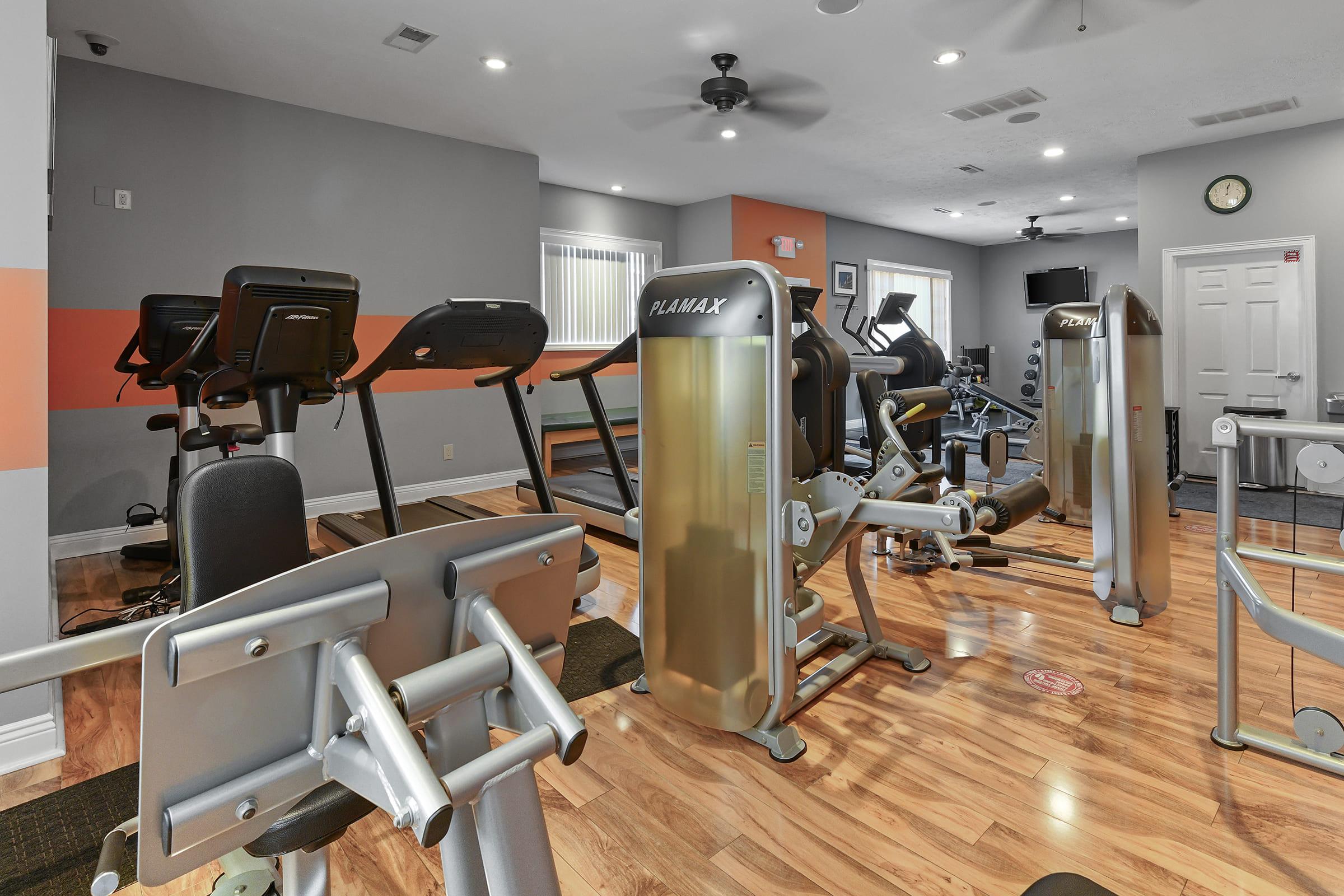 A well-equipped gym featuring various fitness machines, including treadmills, exercise bikes, and weight training equipment. The interior has wood flooring and is lit with natural light. There are wall mirrors and a clock visible, creating a modern workout space.
