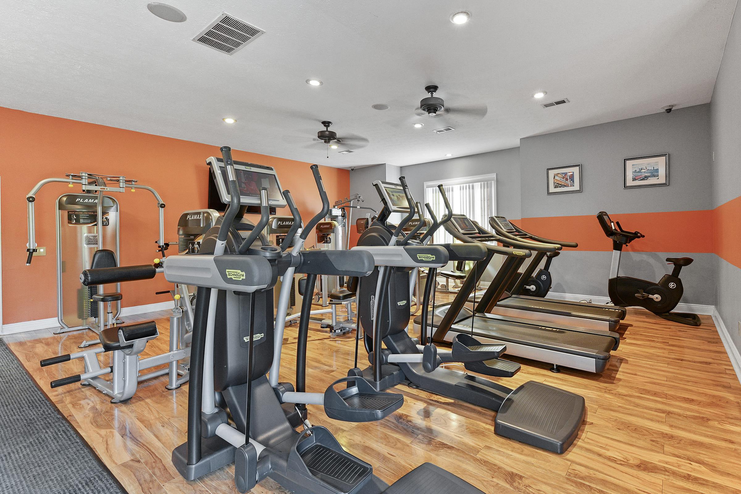 A modern gym interior featuring various exercise equipment, including treadmills, elliptical machines, and weight training machines. The room has orange and gray accents, wooden flooring, and bright overhead lights, creating a spacious and inviting workout environment.