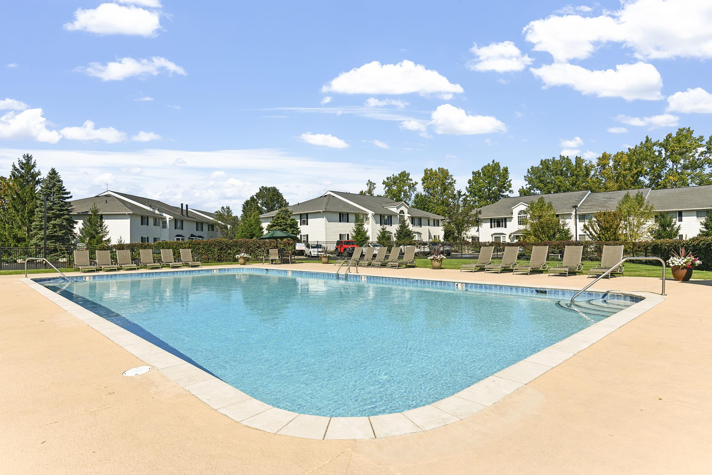 A clear swimming pool surrounded by lounge chairs, with a grassy area and trees in the background. Residential buildings are visible in the distance under a bright blue sky with fluffy clouds.