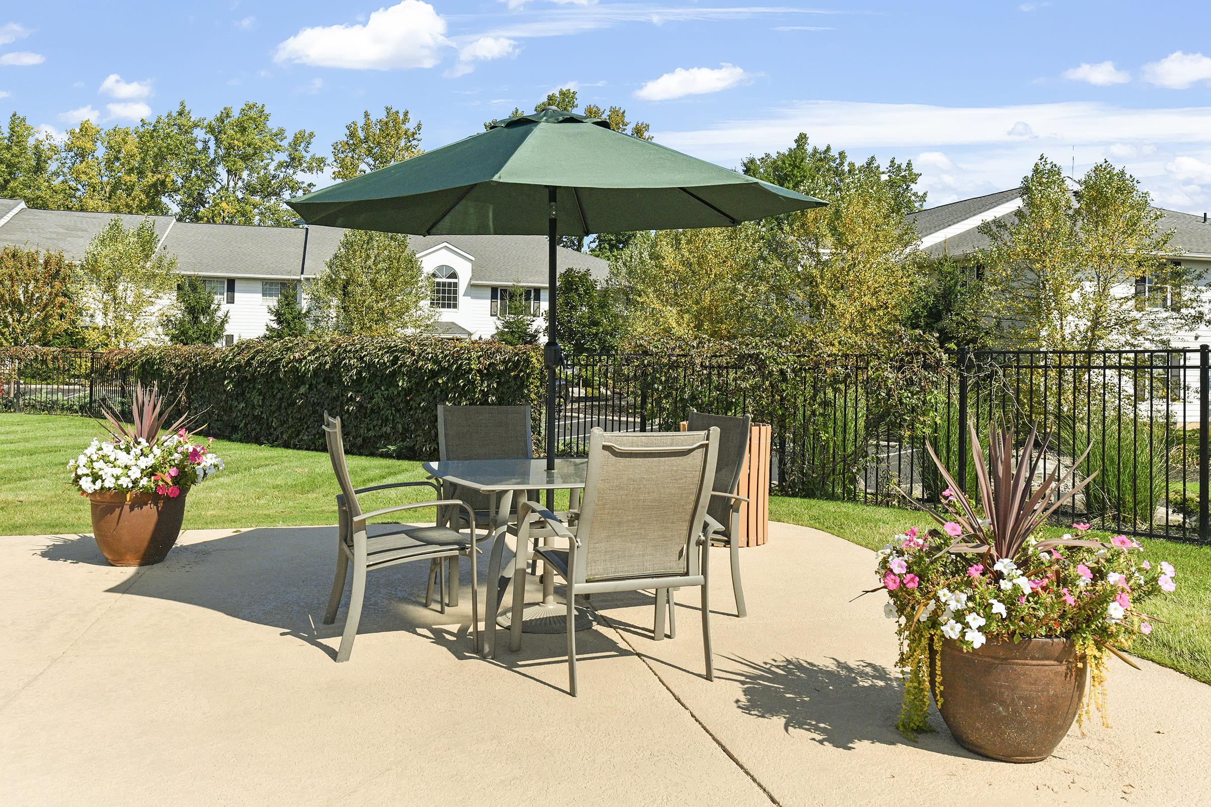 A sunny outdoor patio featuring a green umbrella over a metal table with four chairs. Two large flower pots are adorned with colorful plants on either side. Lush green grass surrounds the area, and a residential building is visible in the background, partially obscured by trees and hedges.