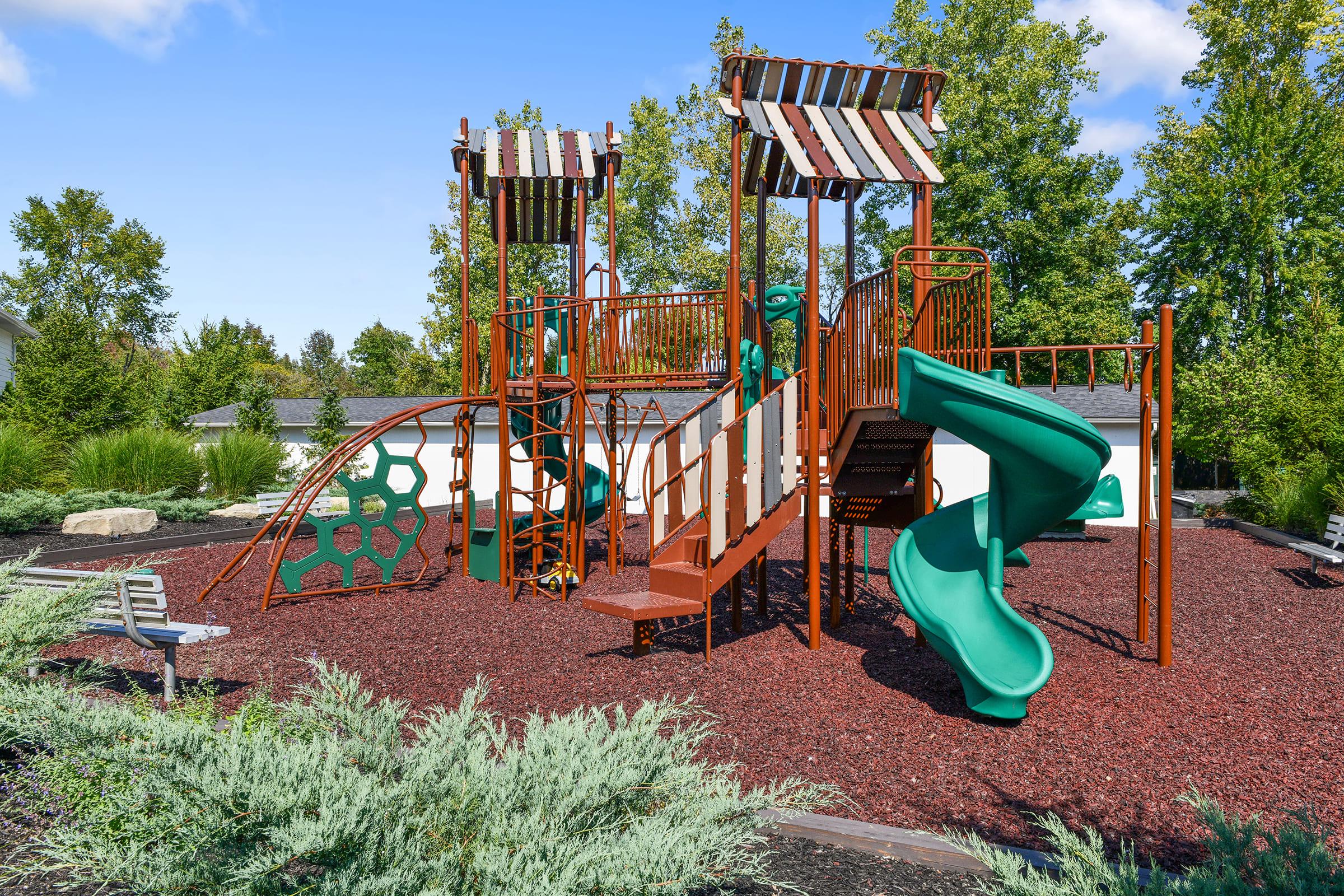 Colorful playground equipment featuring two towers with roofs, a winding green slide, and climbing structures, surrounded by mulch. Lush greenery and trees provide a scenic background, creating an inviting outdoor play area for children.