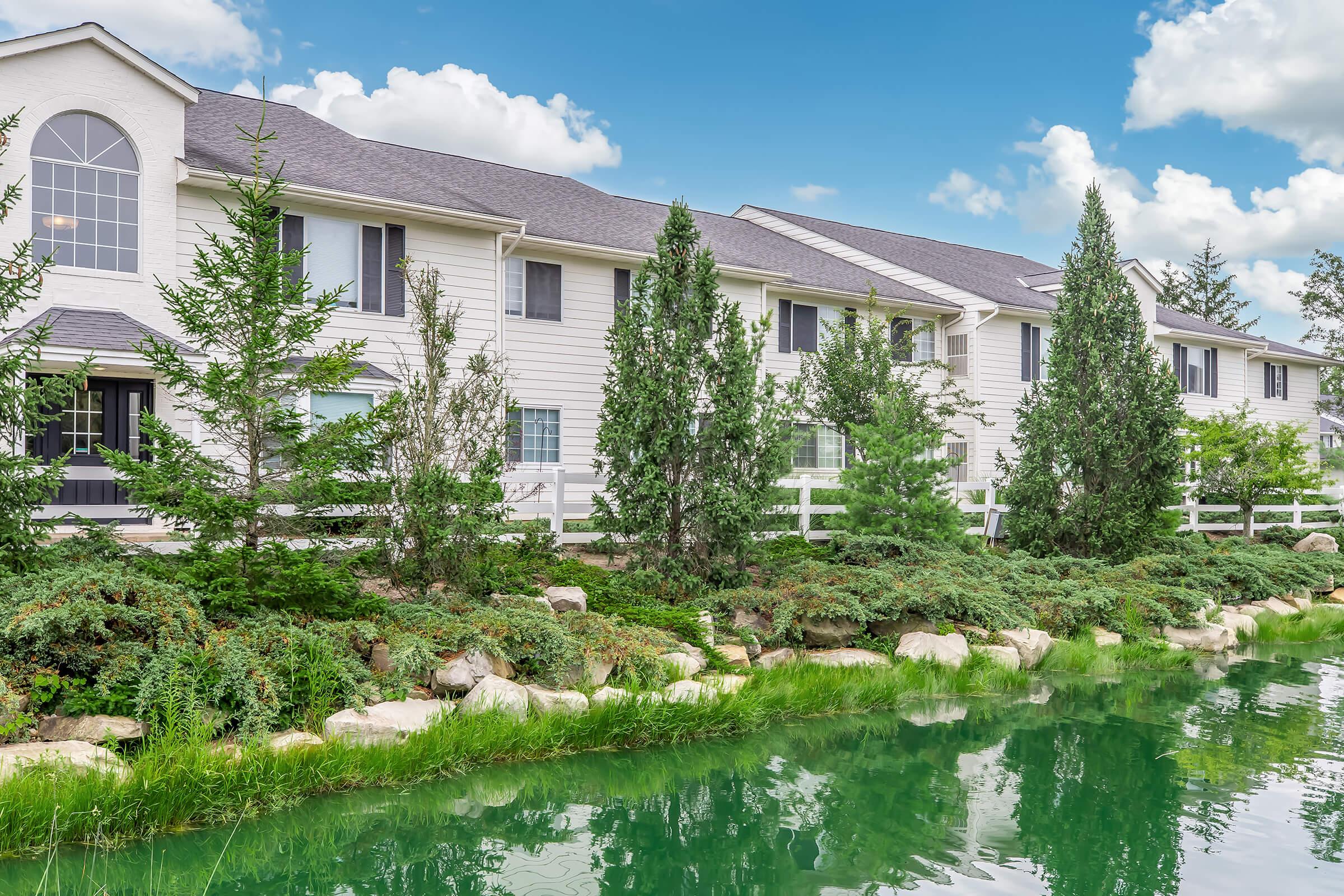 A scenic view of a residential building with light-colored siding, surrounded by lush greenery and ornamental trees. A calm, green pond reflects the sky, and a white fence runs along the property, enhancing the tranquil atmosphere. Fluffy clouds drift in a blue sky above.