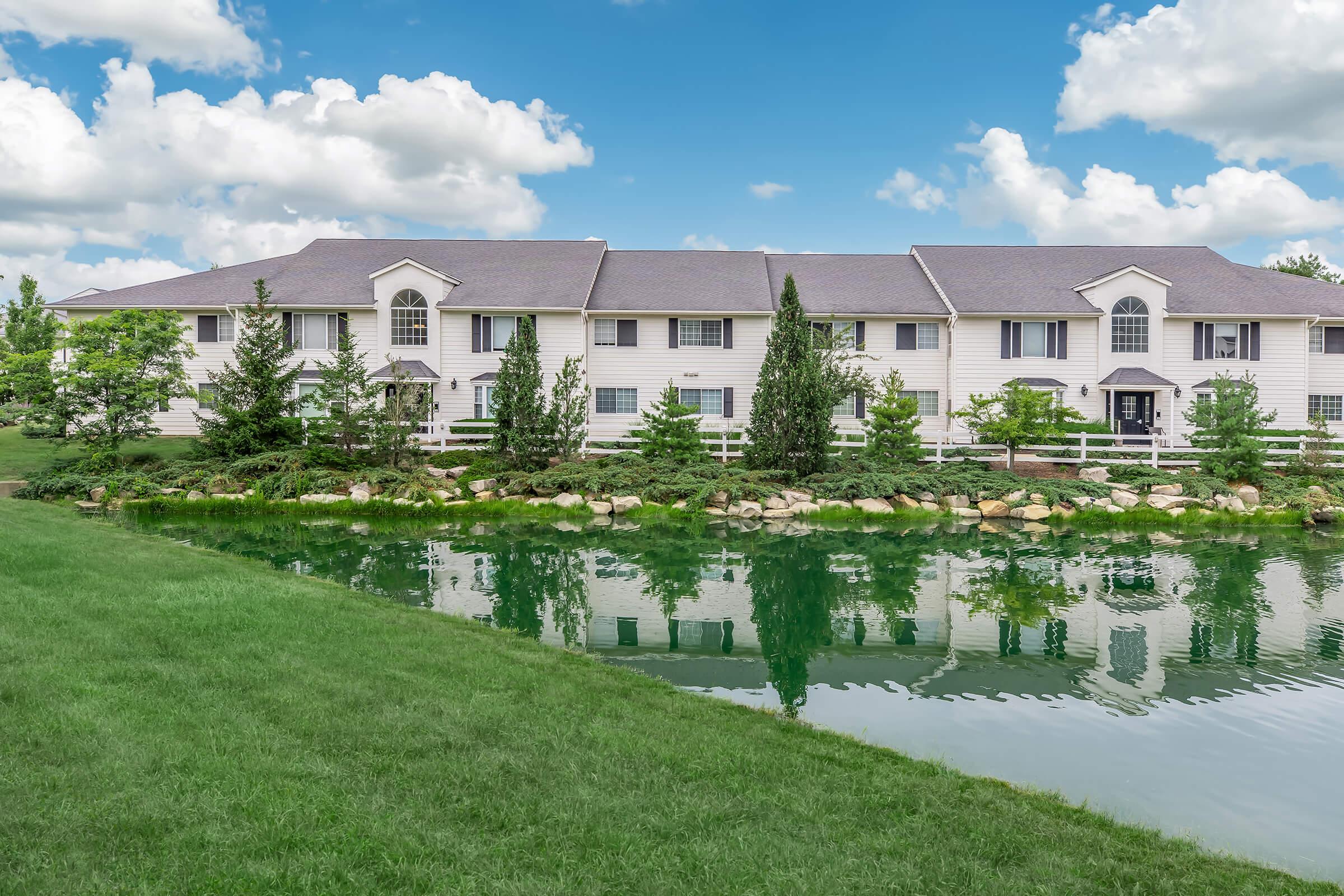 A peaceful residential building with white siding, surrounded by lush green lawns and trees. A pond in the foreground reflects the structure and the blue sky with clouds, creating a serene atmosphere. The scene conveys a sense of tranquility and nature.