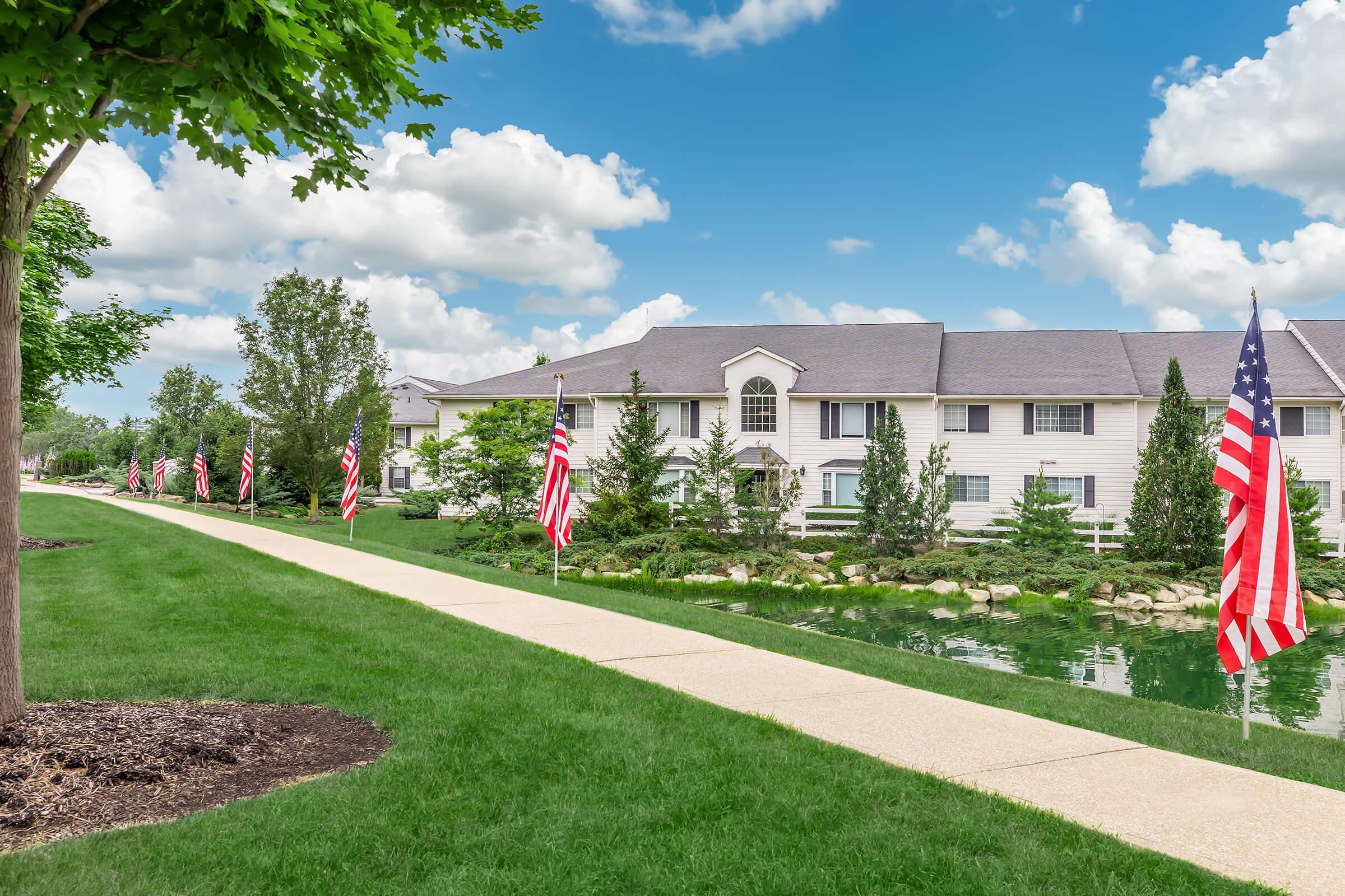 A landscaped pathway lined with small American flags leads to a light-colored building surrounded by greenery and a pond. The sky is bright with fluffy clouds, creating a serene outdoor atmosphere.