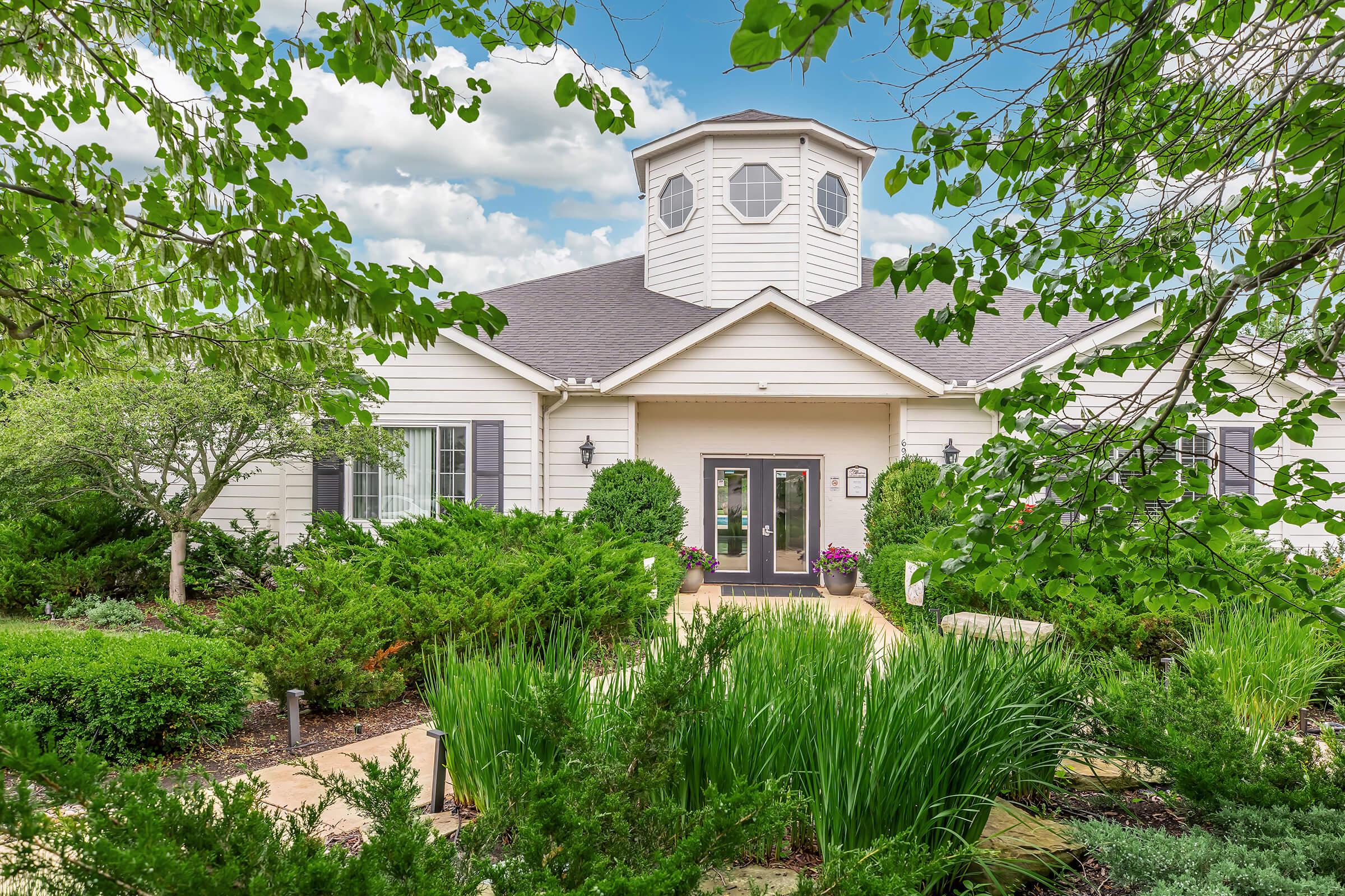 A welcoming suburban home with a round tower feature, surrounded by lush greenery and colorful plants. The entrance has double doors framed by windows, and a stone pathway leads through a landscaped garden. The sky is bright with fluffy clouds, creating a picturesque setting.