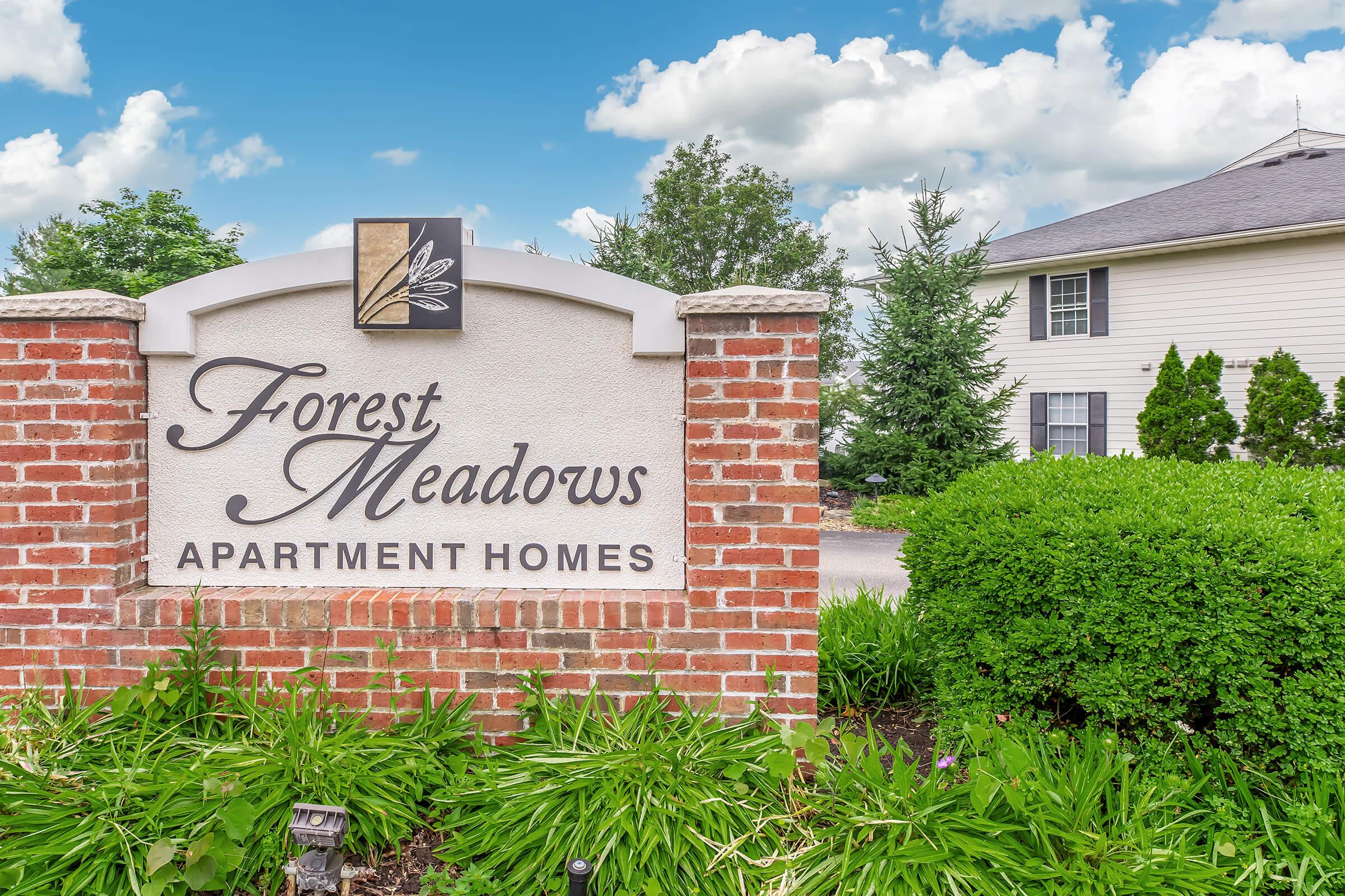 Sign for Forest Meadows Apartment Homes displayed on a brick pillar, surrounded by lush green landscaping. In the background, a residential building is partially visible under a bright blue sky with fluffy white clouds.