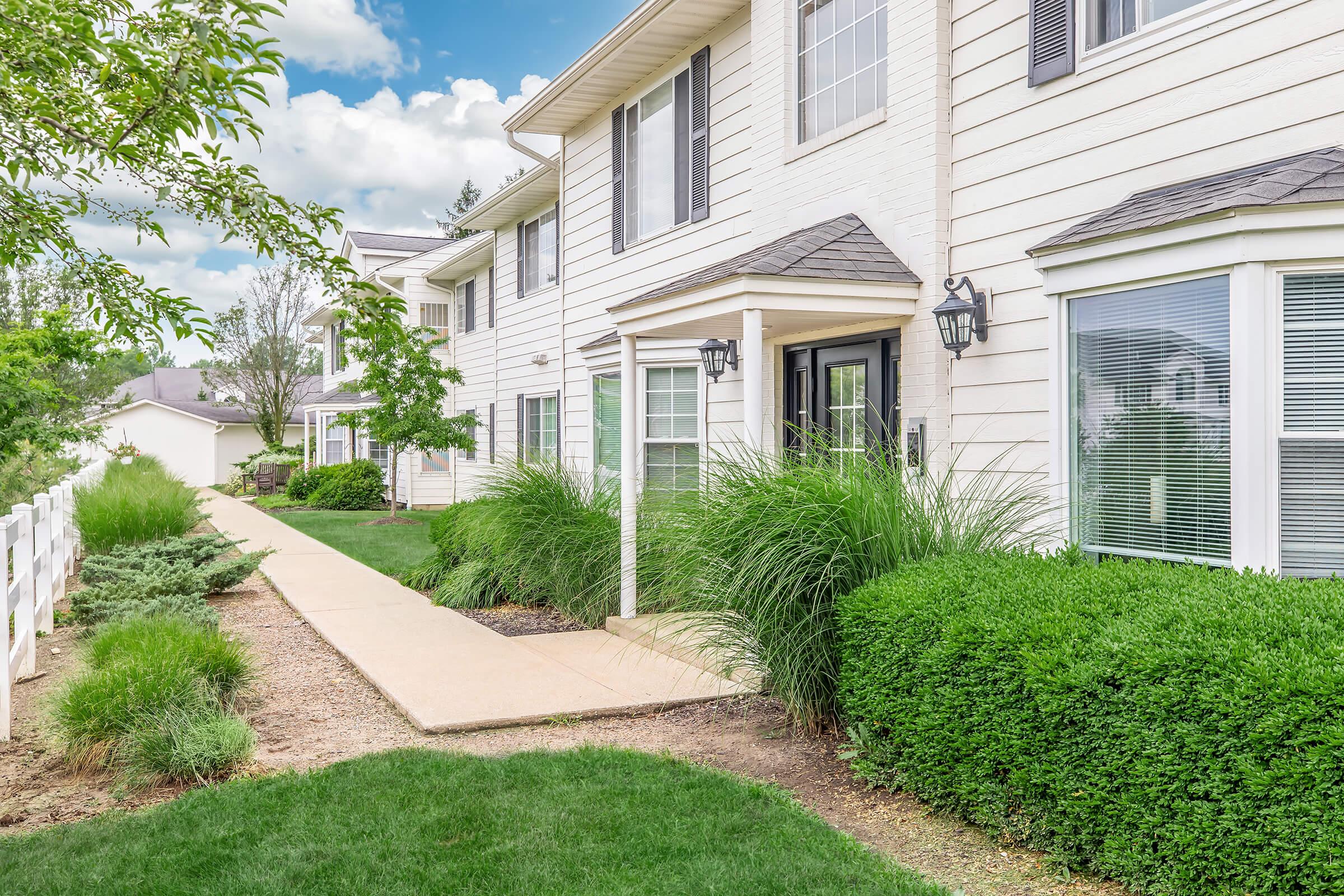 A well-maintained residential building with white exterior walls, featuring large windows and a welcoming entrance. Lush green grass and neatly trimmed bushes line the pathway leading to the front door, with trees and shrubs enhancing the landscaped surroundings under a partly cloudy sky.