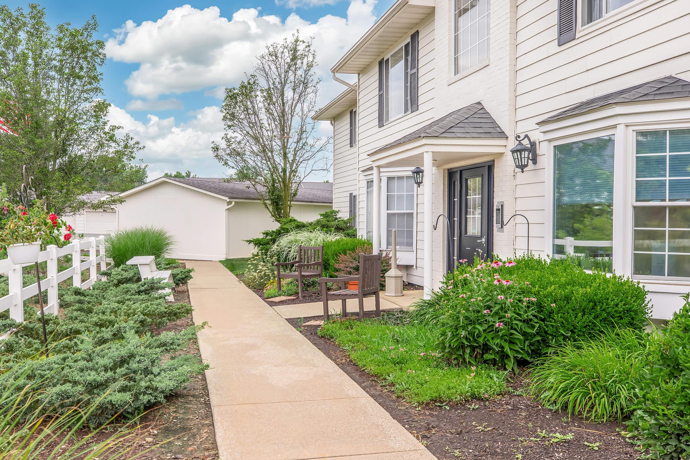 A charming suburban home with a well-maintained front yard featuring neatly trimmed bushes and flowers. A concrete pathway leads to the front door, flanked by wooden benches. Lush greenery and a white fence add to the inviting atmosphere, under a partly cloudy sky.