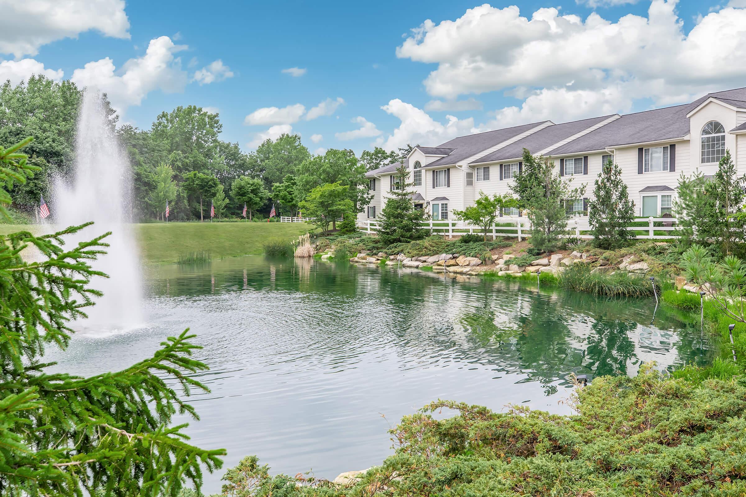 A tranquil scene featuring a pond with a fountain, surrounded by lush greenery and trees. In the background, there are white buildings with balconies, and a clear blue sky dotted with fluffy clouds reflects in the water. The area is beautifully landscaped, creating a peaceful atmosphere.