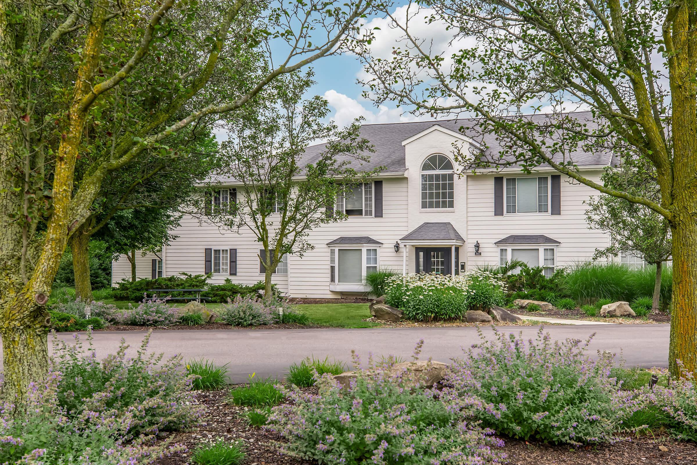 A large, modern two-story house with white siding and gray roof, surrounded by lush greenery and flowering plants. The driveway leads up to the front entrance, framed by trees. The sky is partly cloudy, creating a serene and inviting atmosphere.