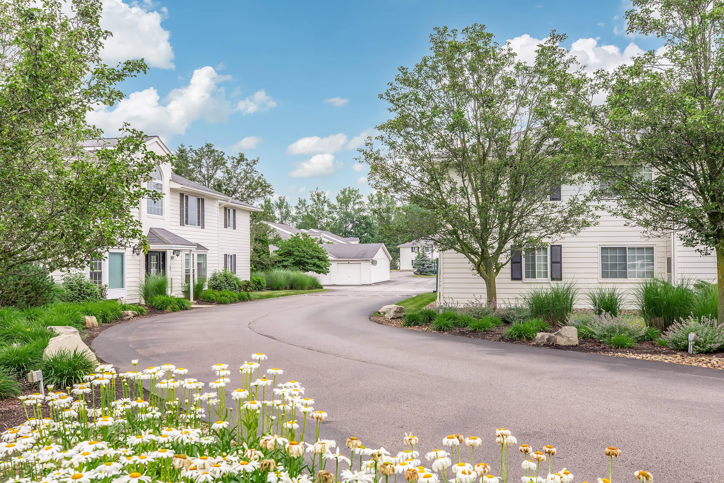 A serene residential neighborhood featuring white two-story houses surrounded by lush green trees and vibrant flowers. A winding paved road meanders through the area, leading to a row of garages in the background under a bright, partly cloudy sky.