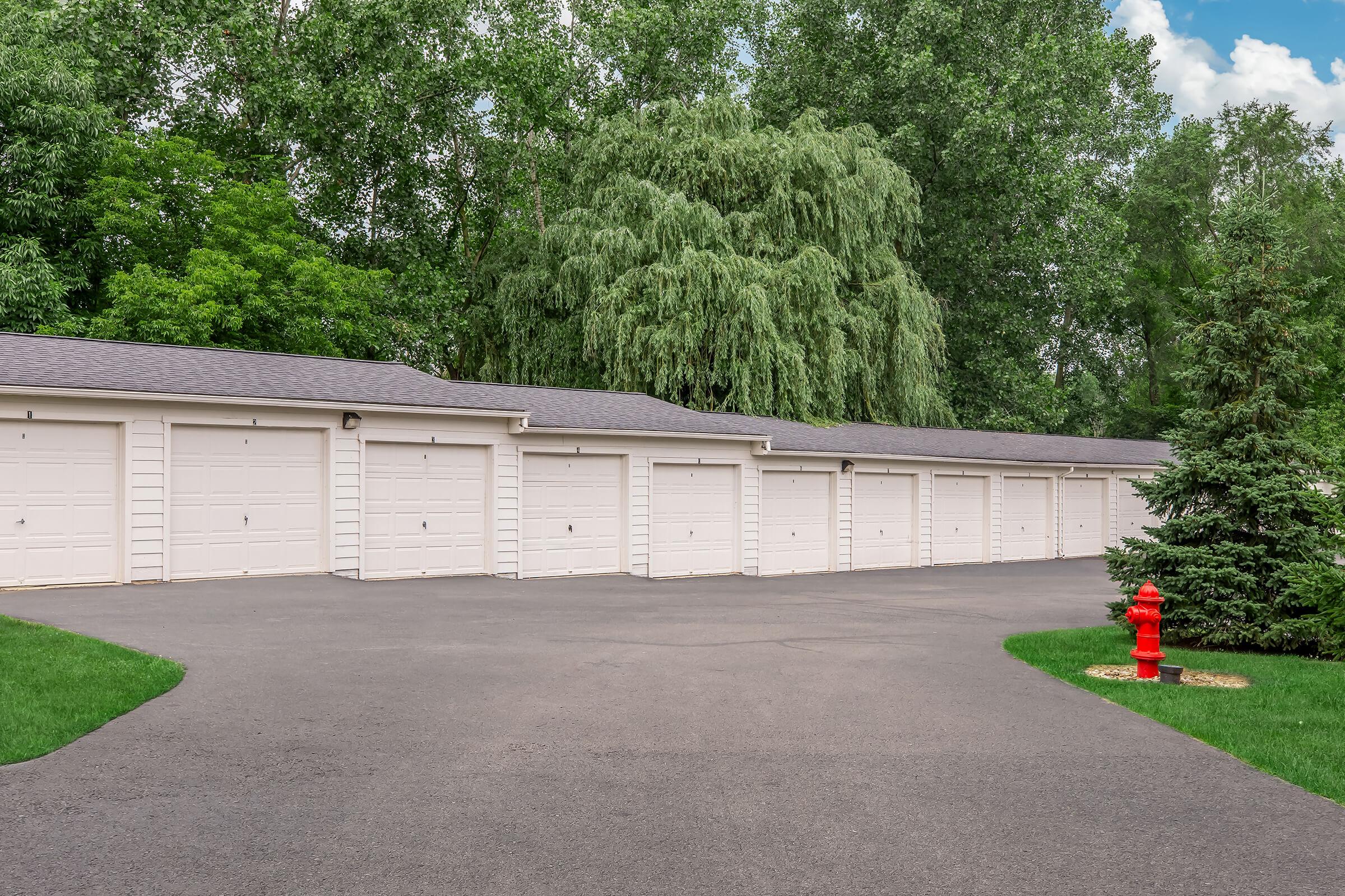 Row of white garage doors lined up along a paved driveway, surrounded by lush green trees. A red fire hydrant sits near the edge of the driveway, adding a pop of color to the serene landscape. The scene is bright and inviting.