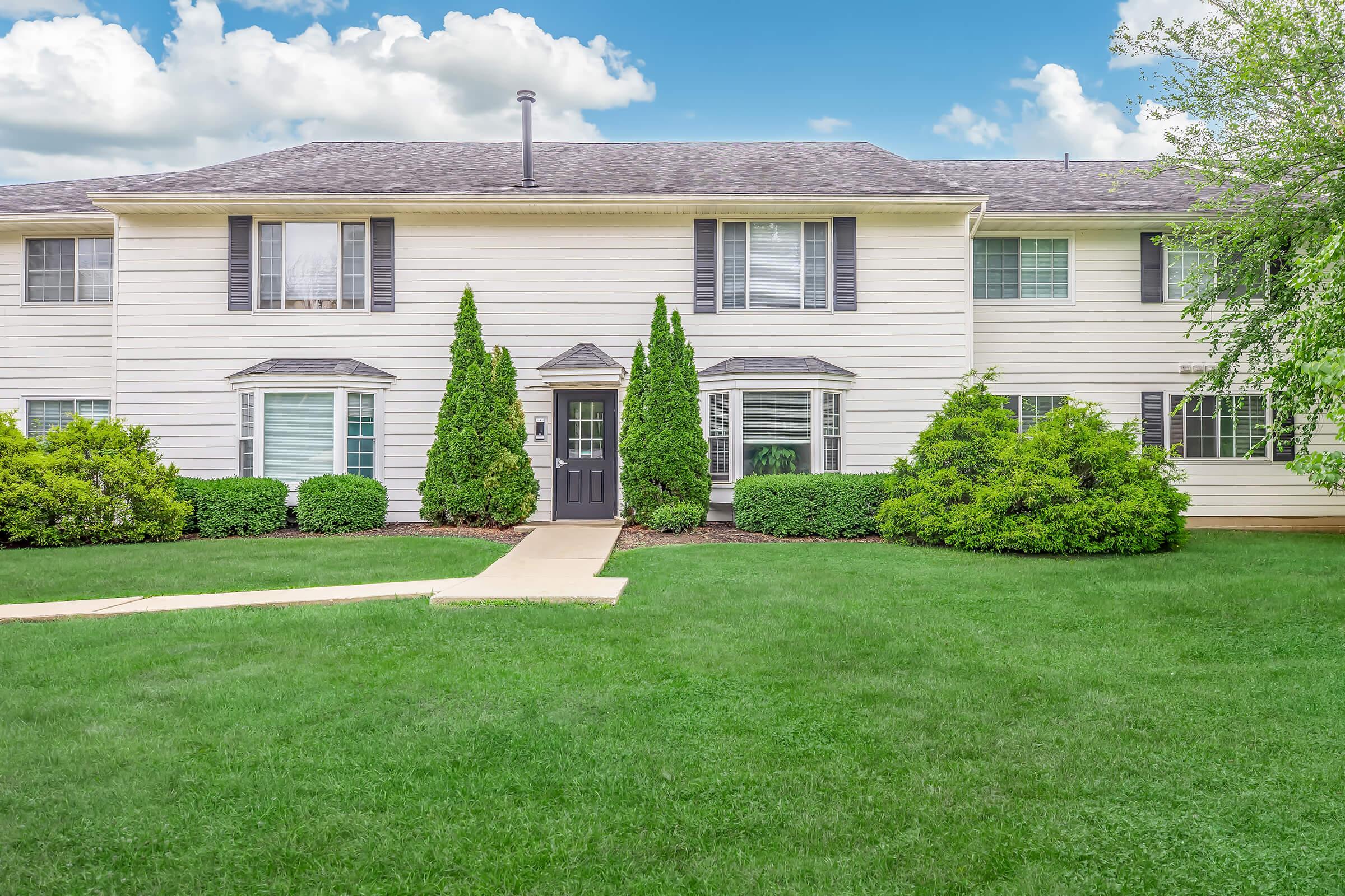A two-story residential building with white siding and multiple windows. The entrance features a dark door surrounded by green shrubs and small trees. A neat lawn with well-maintained grass is in front of the building, and the sky above is partly cloudy.