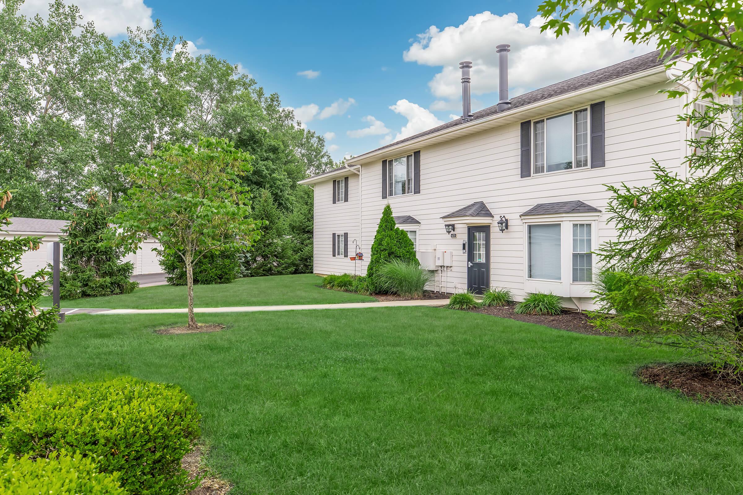 A well-maintained two-story residential building with light siding, surrounded by lush green grass and trees. The front yard features manicured shrubs and plants, and a paved walkway leading to the entrance. White clouds can be seen in the blue sky above.