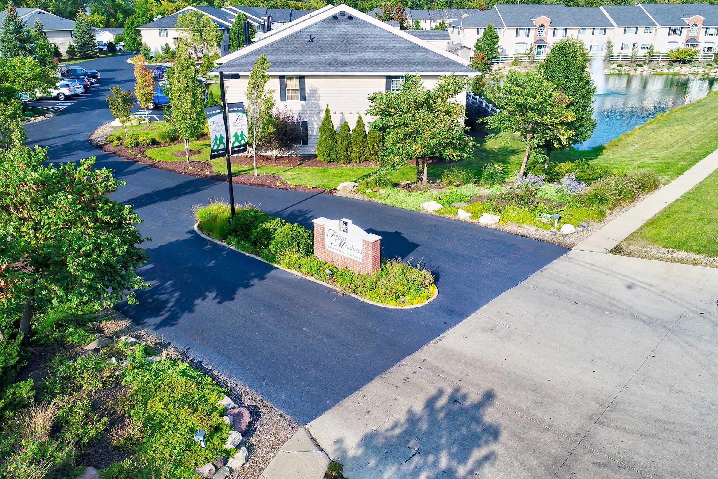 An aerial view of a residential community entrance featuring a sign that reads "The Meadows." The area includes well-maintained landscaping, trees, and a small pond nearby. The road is lined with parked cars, and the surrounding buildings are two-story residences.