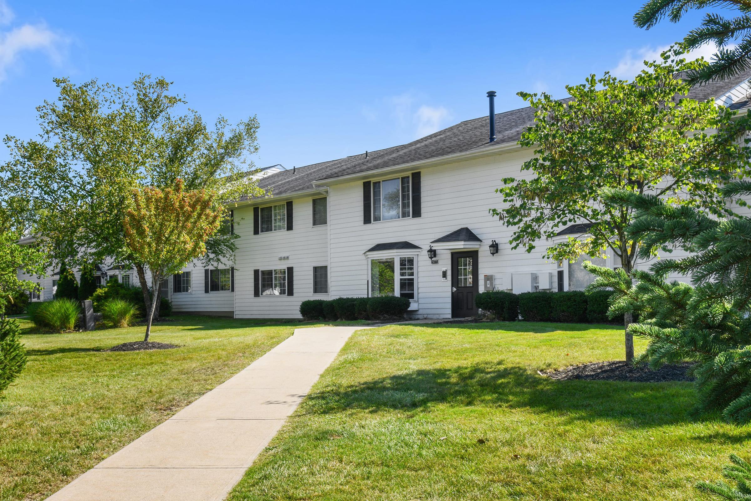 A well-maintained, two-story apartment building with white siding, black shutters, and a concrete pathway leading to the entrance. Lush green grass and trees are visible in the landscaped yard, under a bright blue sky.