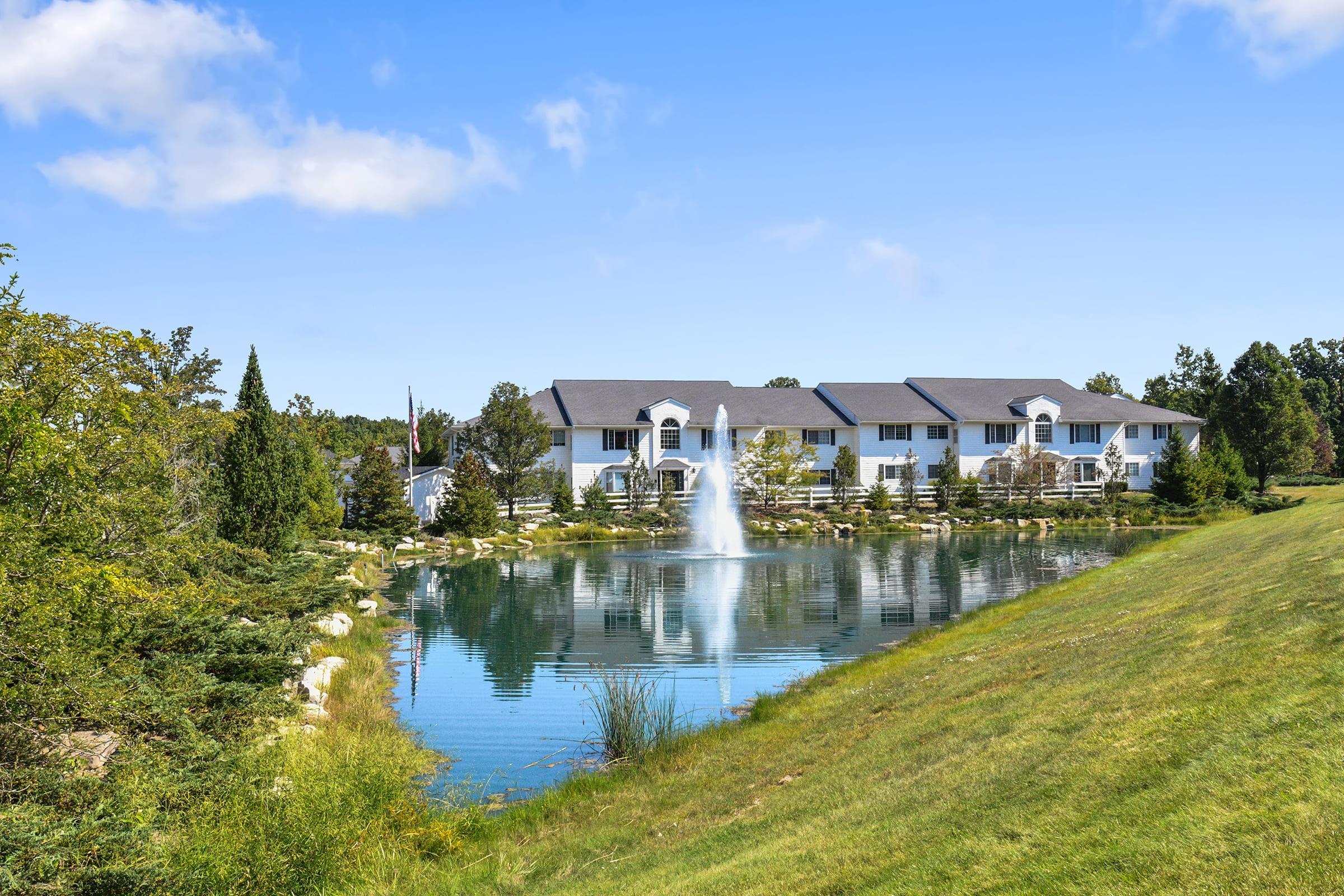 A serene landscape featuring a calm pond with a fountain surrounded by lush greenery. In the background, a well-maintained building with a sloped roof is visible. The scene is set under a clear blue sky, creating a peaceful and inviting atmosphere.