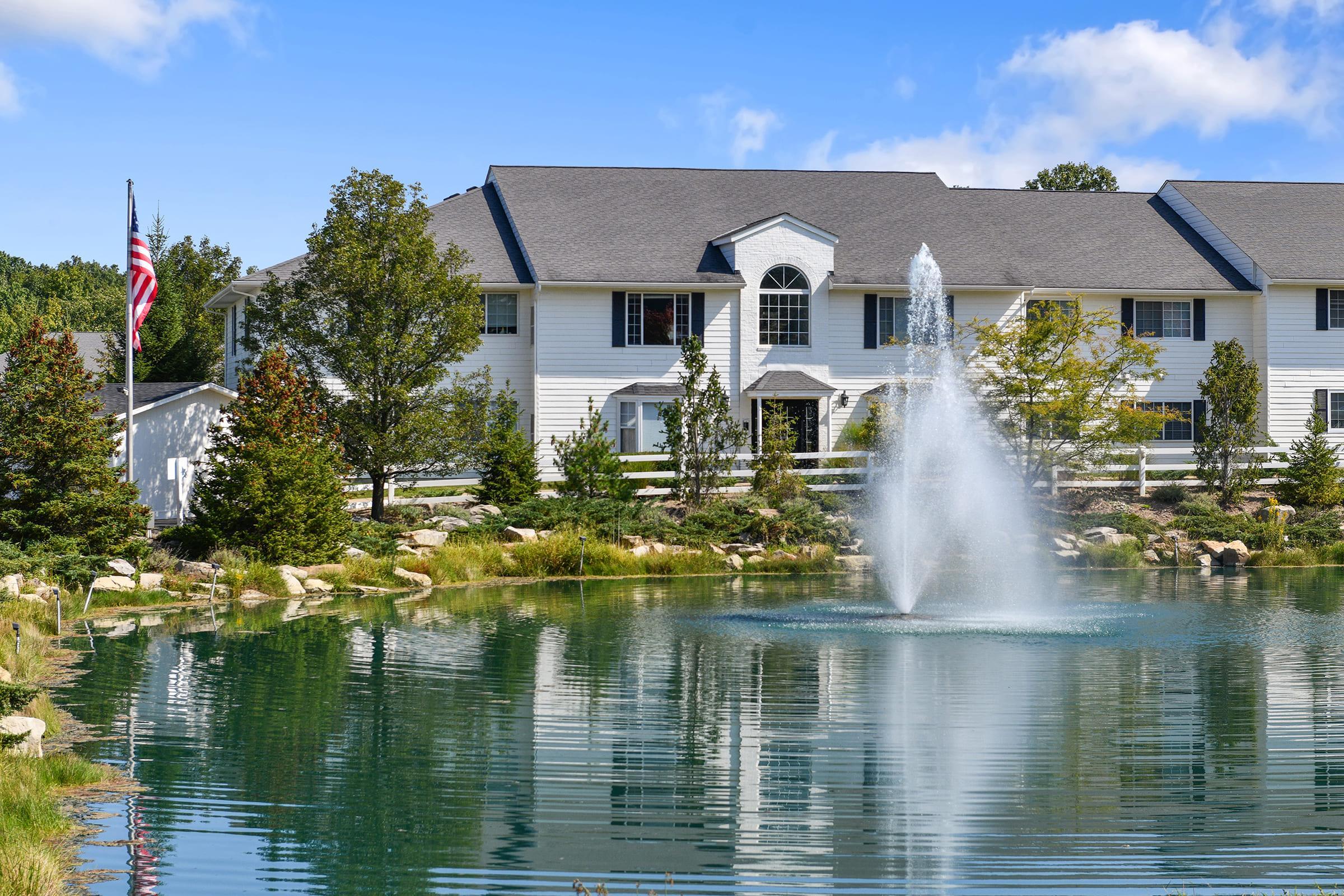 A serene landscape featuring a clear pond with a fountain, surrounded by lush greenery and small trees. In the background, a white multi-story building can be seen, with a flagpole displaying the American flag nearby. The scene is bright and inviting, reflecting a peaceful suburban atmosphere.