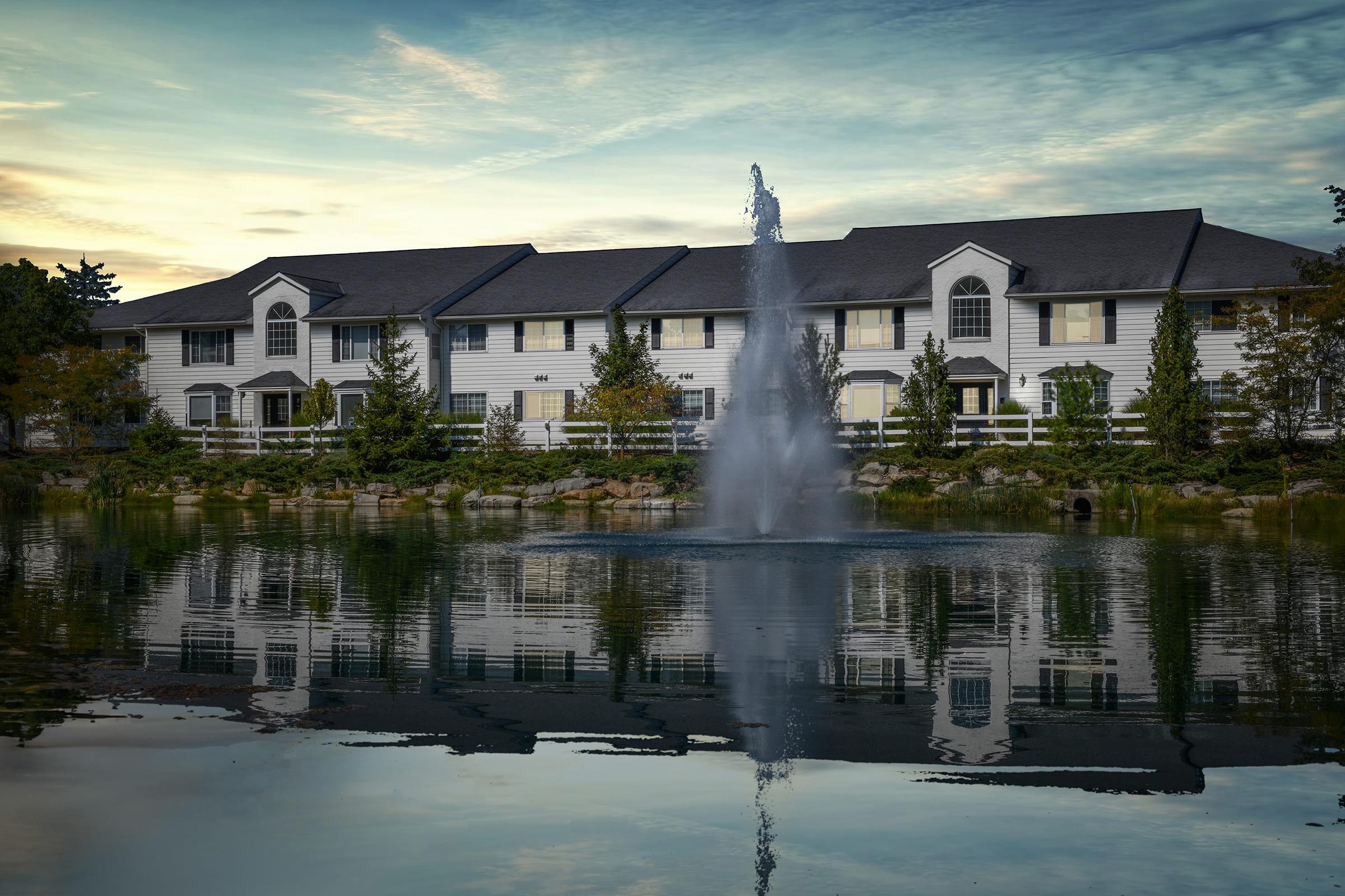 A tranquil scene featuring a charming residential building with a fountain in front, surrounded by lush greenery. The building reflects in the calm water of the pond, under a cloudy sky at sunset, creating a serene and picturesque atmosphere.