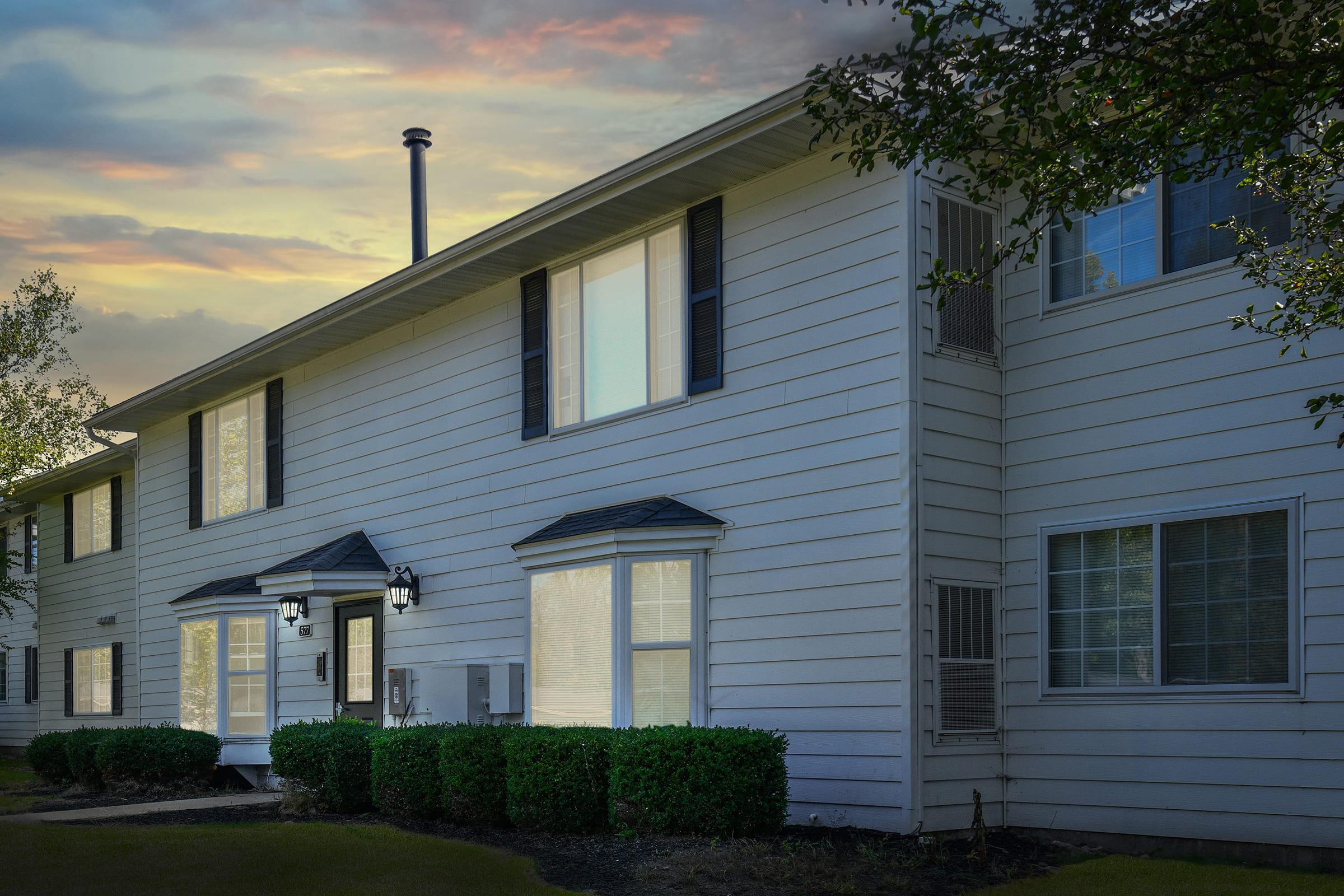 A white two-story residential building with multiple windows and a black roof. The façade features well-manicured hedges in front, and there are outdoor light fixtures beside the entrance. The sky in the background is painted with soft hues, indicating either sunrise or sunset.