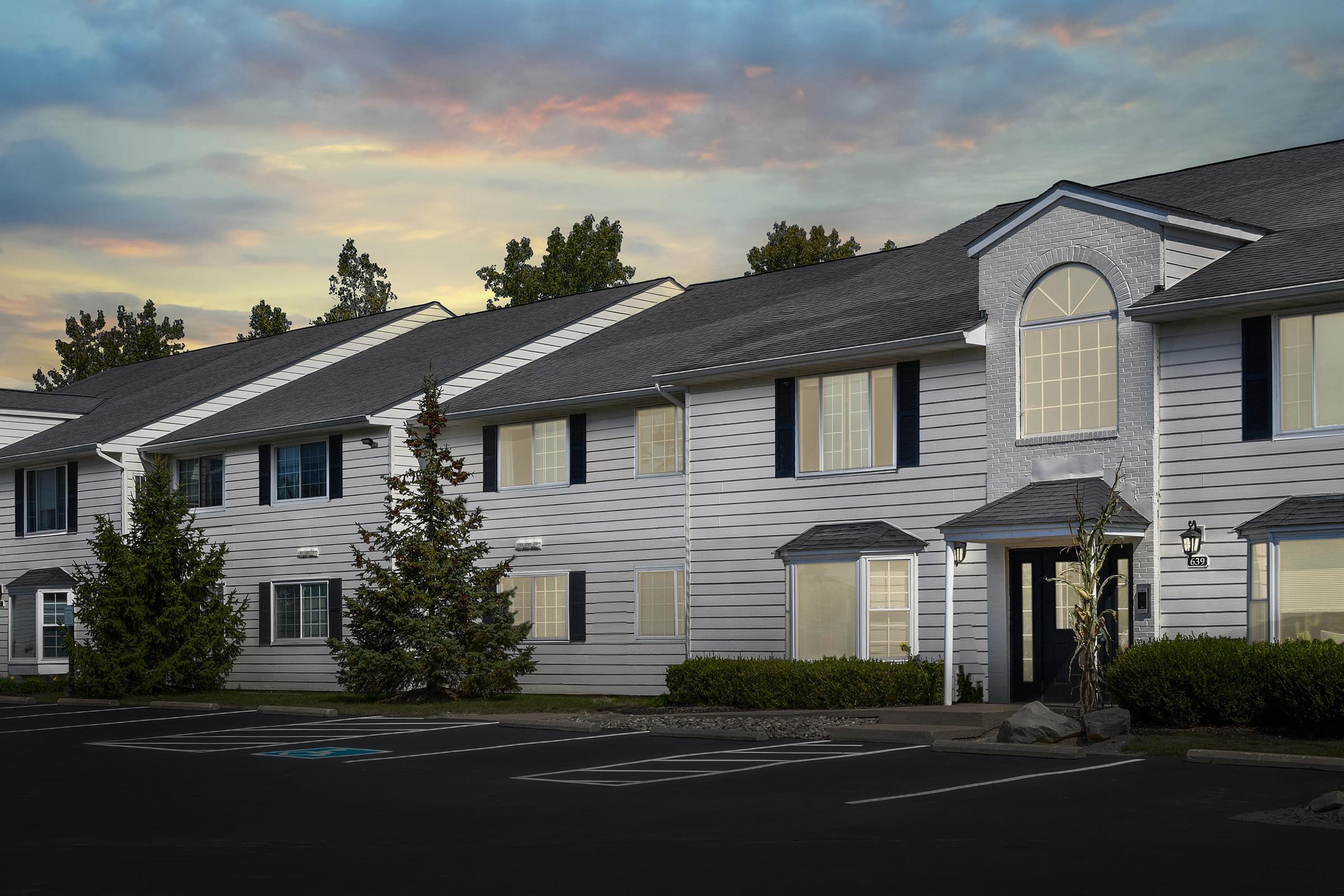 A row of modern apartment buildings with light gray siding, featuring long windows and blue shutters. In the foreground, a parking lot with a few trees and landscaped shrubs. The background displays a colorful sky at dusk, adding a warm ambiance to the scene.