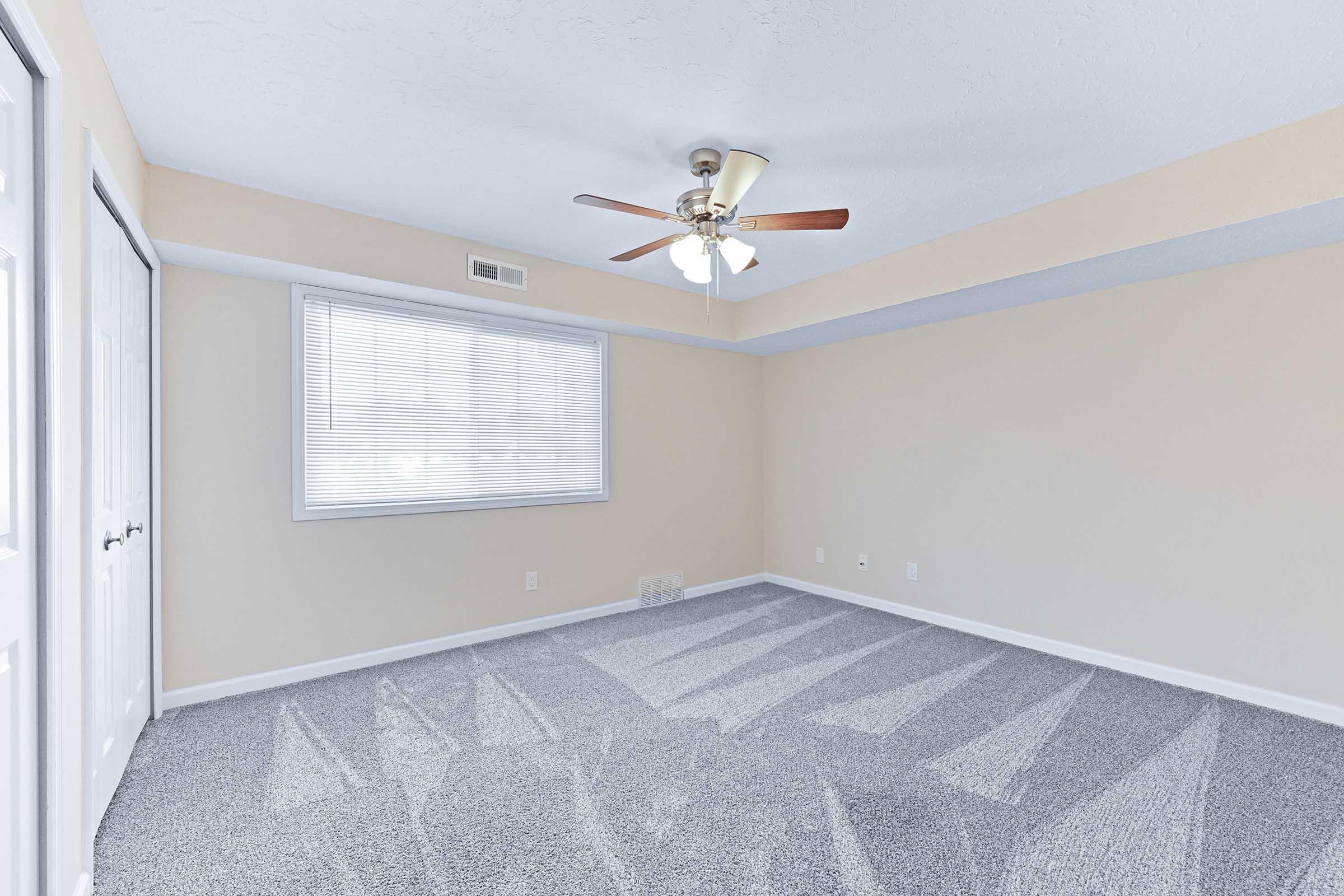 A well-lit empty room featuring light beige walls, a ceiling fan with wooden blades, and a window with blinds. The floor is covered in gray carpeting, and there are two double doors on one side. The room has a clean and spacious appearance, ideal for various uses.