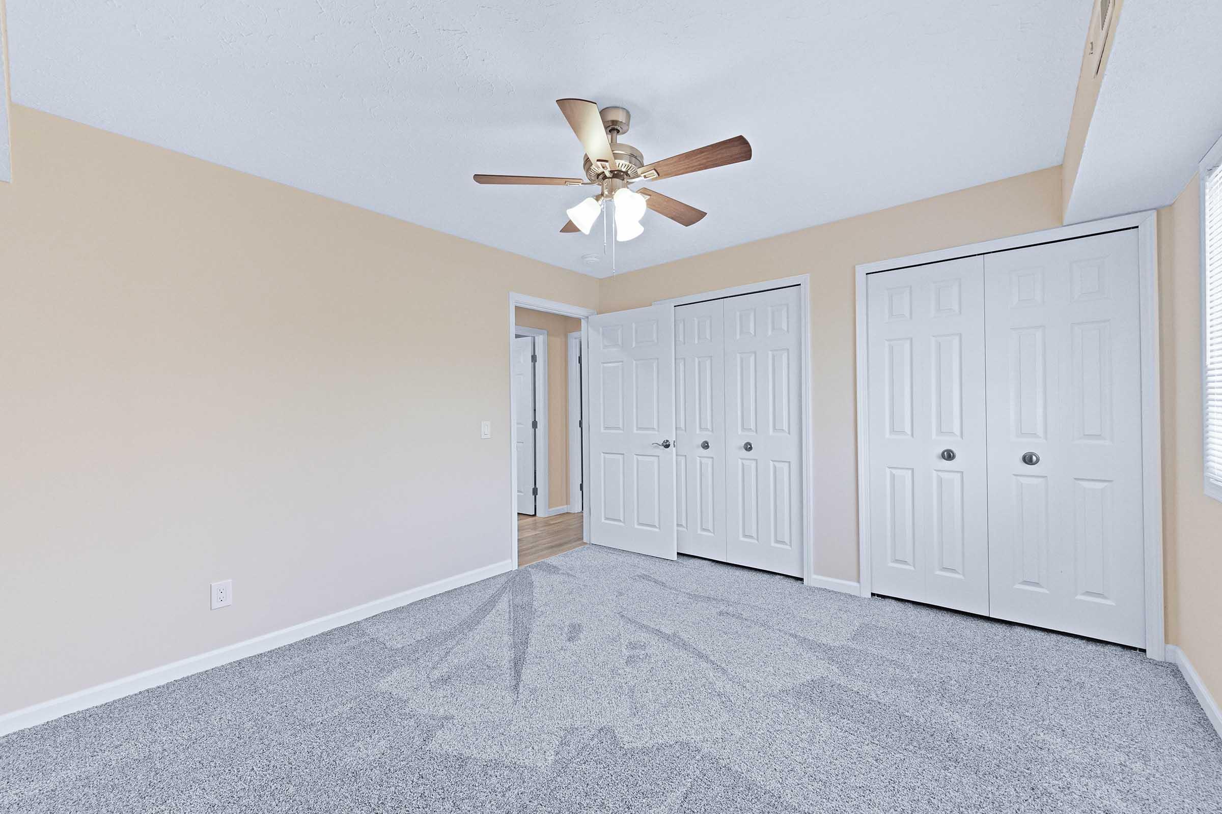 A spacious room with light beige walls and gray carpet. There's a ceiling fan with wooden blades, and two white double doors leading to closets. A doorway in the background connects to another room, and natural light streams in through a window, enhancing the airy feel of the space.