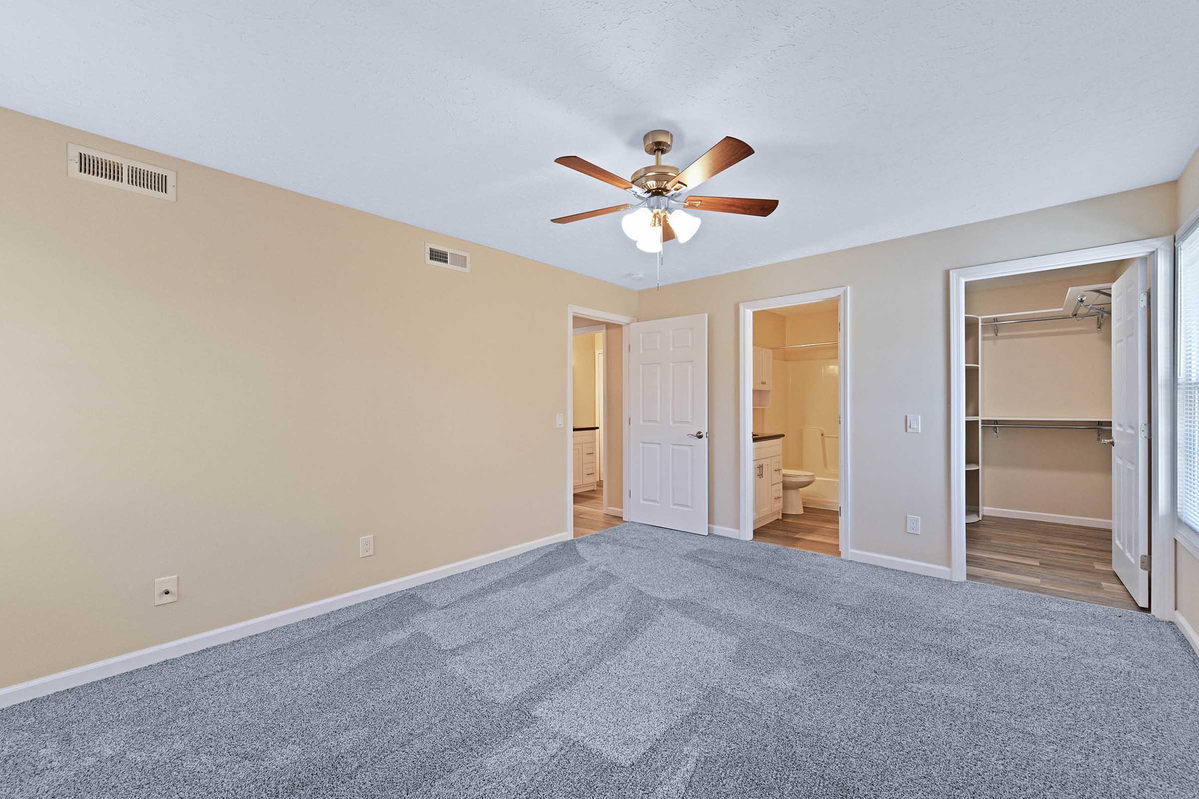 A spacious, well-lit room with light beige walls and a ceiling fan. The floor is covered in plush gray carpeting. Two doorways lead to other areas, one of which opens to a bathroom. A closet with shelving is visible on the right. The overall atmosphere is clean and inviting.