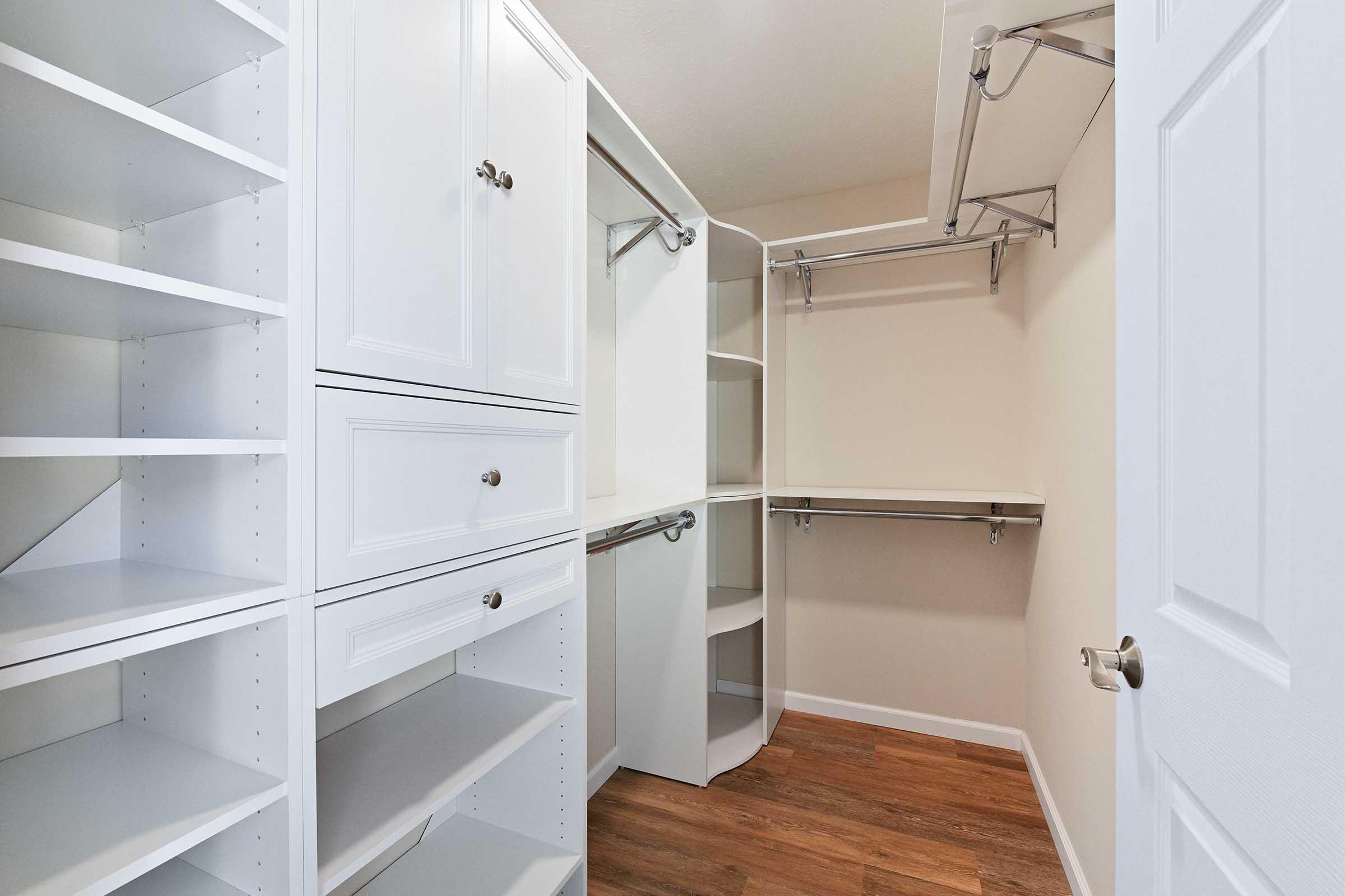 A spacious, well-organized closet featuring white shelving units, hanging rods, and drawers. The closet has a light-colored wall and a warm wooden floor, creating a bright and inviting storage space. The design allows for efficient organization of clothes and accessories.