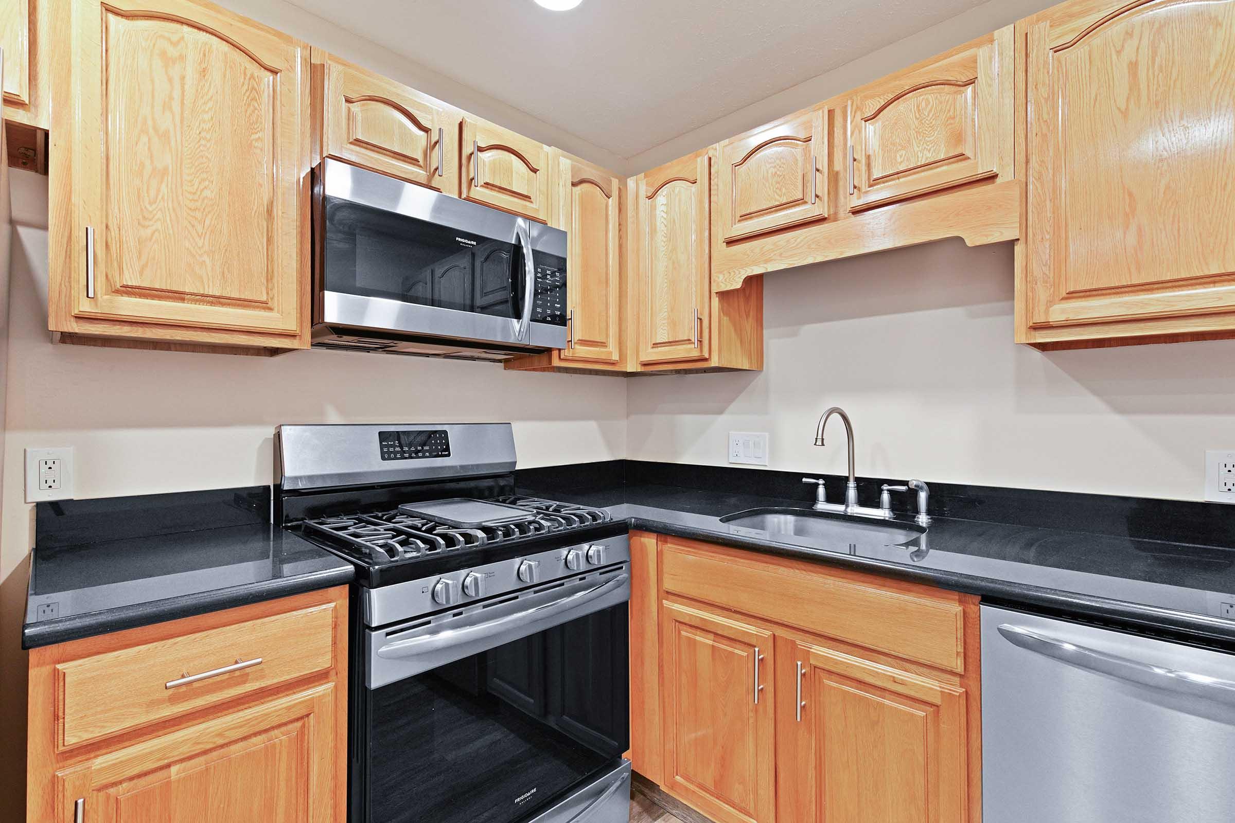 Modern kitchen featuring wooden cabinets, stainless steel appliances, a gas stove, and a sleek black countertop. The sink is situated next to the stove, with a silver dishwasher visible to the right. Warm lighting enhances the inviting atmosphere of the space.