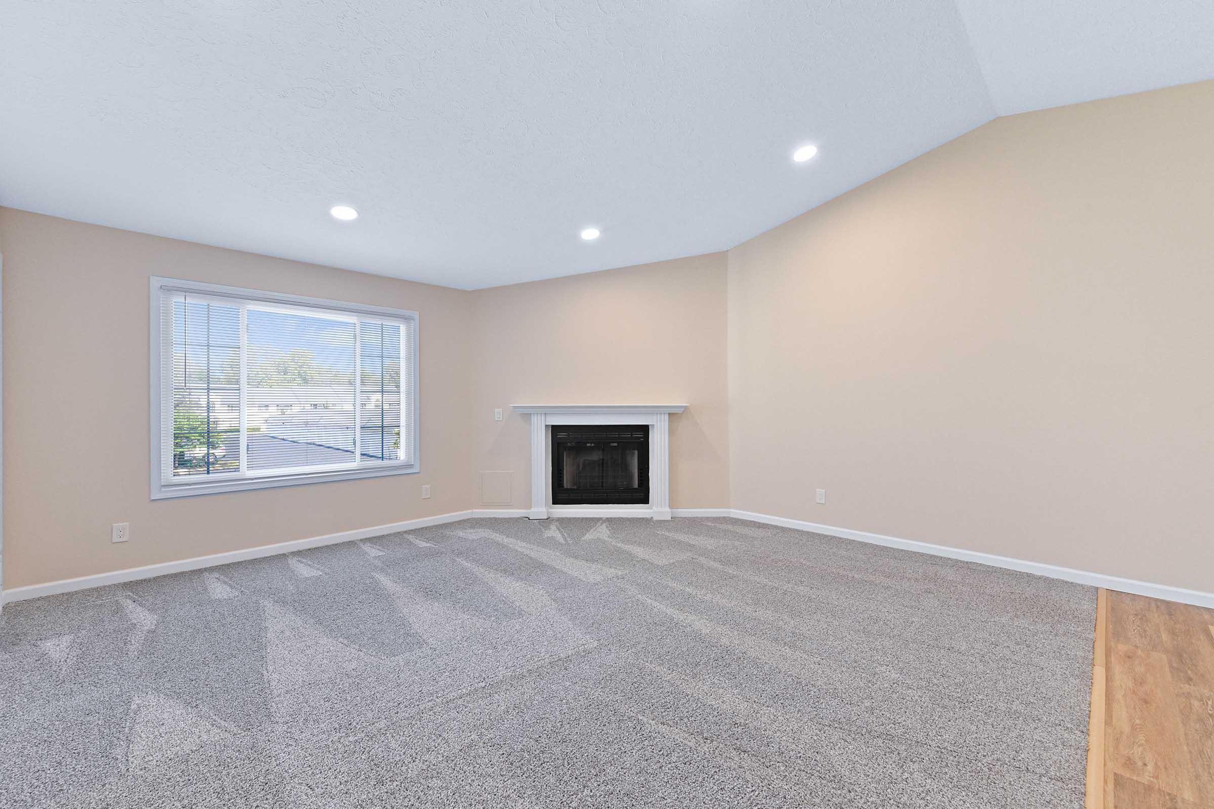 A spacious, well-lit living room with light beige walls and a cozy fireplace. Large windows allow natural light to flow in, highlighting the soft gray carpet. The ceiling is vaulted, creating an open and airy atmosphere, while the room is free of furniture, offering a blank canvas for decorating.