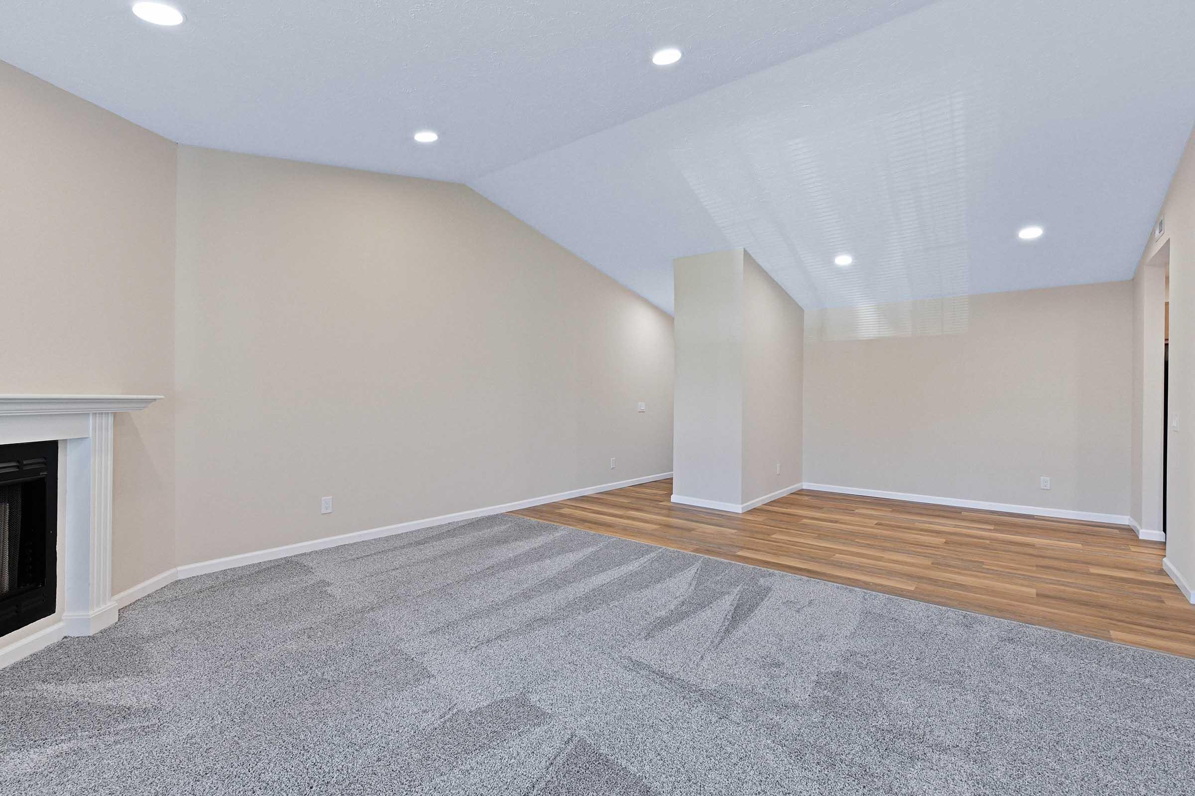 Interior of a spacious room featuring light-colored walls and a vaulted ceiling. The flooring consists of a blend of carpet and hardwood. There is a fireplace on the left side, and the room is well-lit with recessed lighting. The open space is empty, allowing for flexible furniture arrangement.