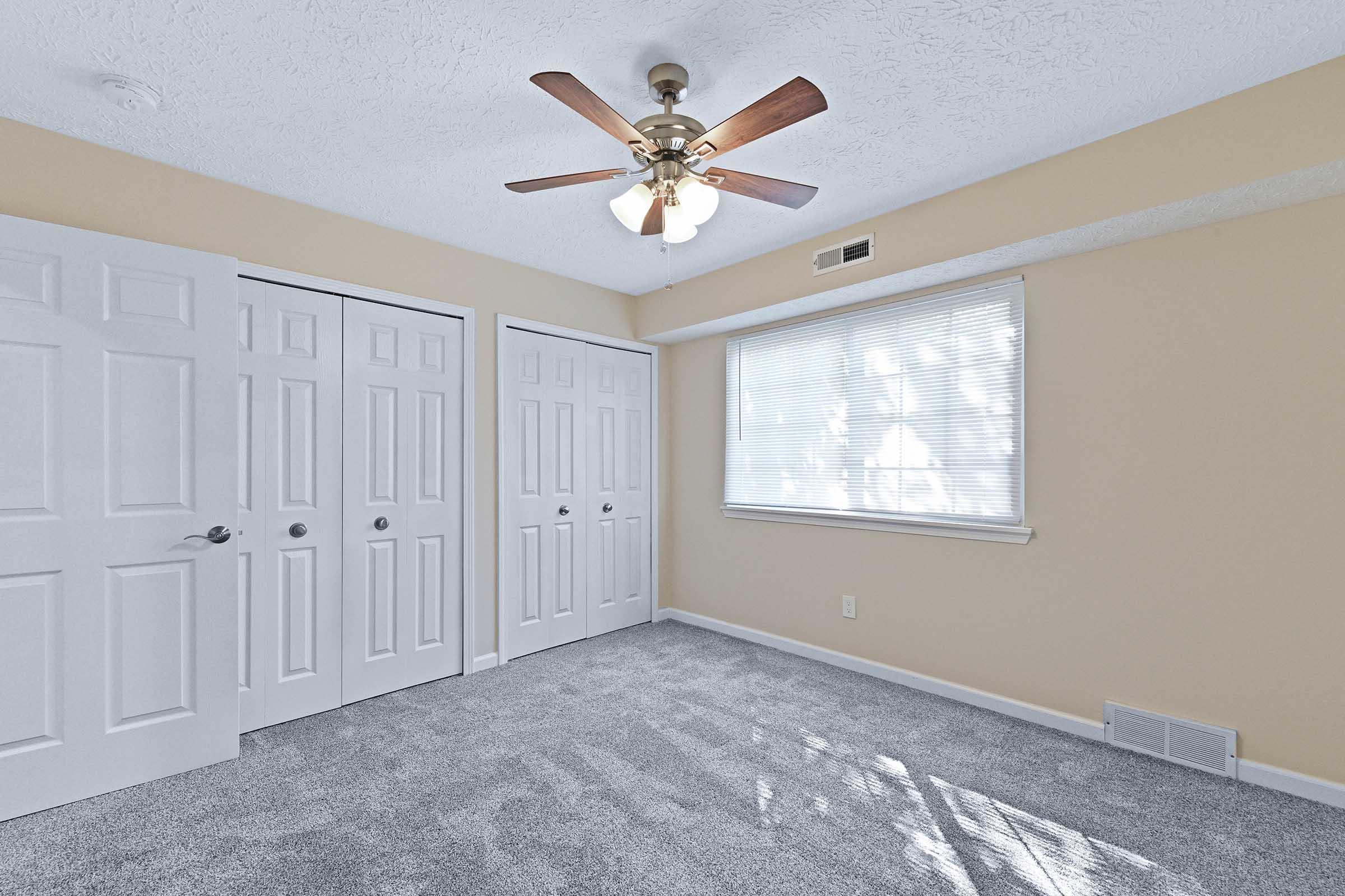A well-lit bedroom featuring light beige walls, a ceiling fan with light, and plush gray carpet. There are two white double doors leading to closets, and a window with sheer curtains allowing natural light to enter the room.