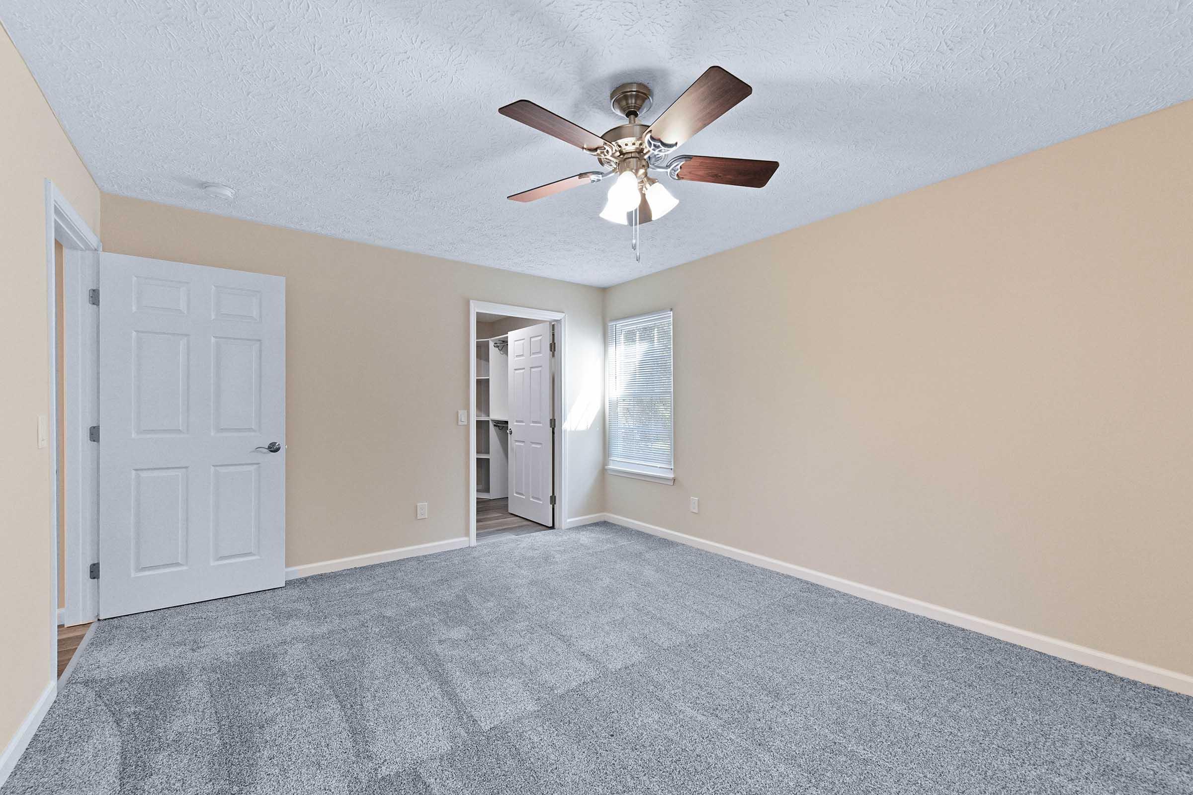 Spacious room with light beige walls and gray carpet. A ceiling fan with wooden blades hangs above. There are two white doors: one leading to a closet and the other to another room. A window allows natural light to enter, creating a bright and airy atmosphere. Ideal for various uses like a bedroom or office.