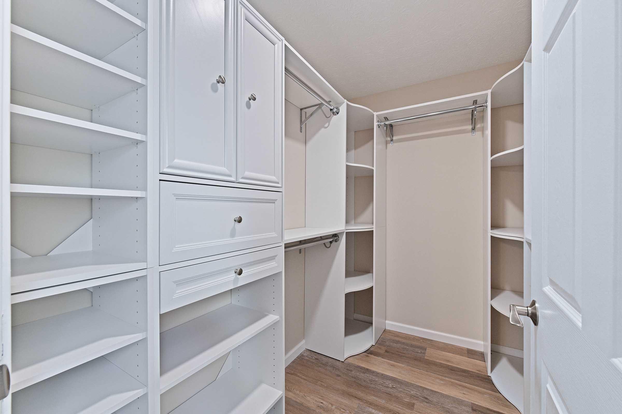 A spacious, organized closet featuring white shelving units, drawers, and a hanging rod. The closet has a light beige wall and wood laminate flooring, creating a clean and modern storage area.