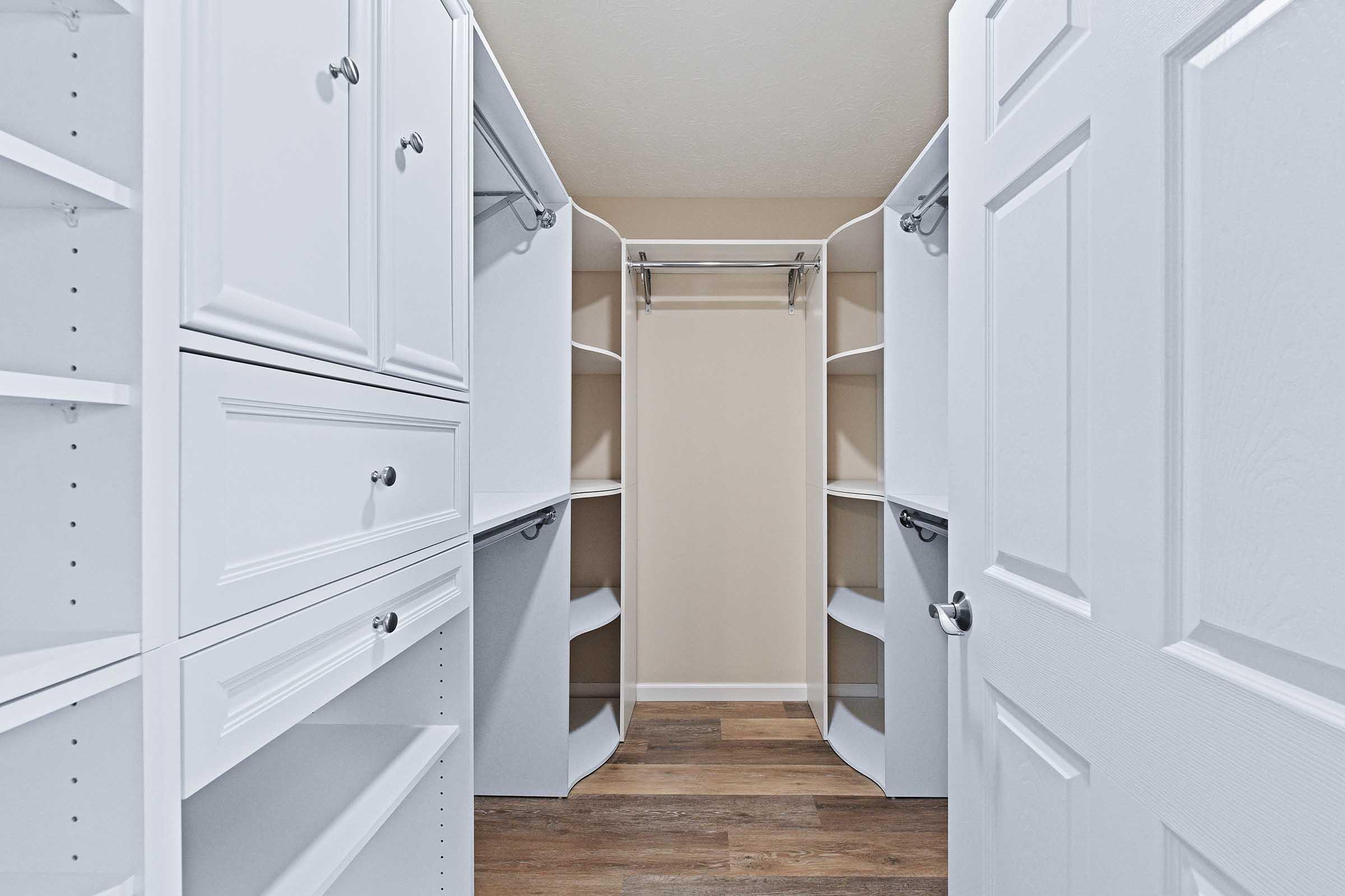 A spacious walk-in closet featuring white shelving and drawers. The room has two open sides with shelving units, and a central hanging rod. The flooring is wooden, and the walls are a neutral color, adding to the overall bright and organized appearance of the space. A white door is slightly ajar on the left.