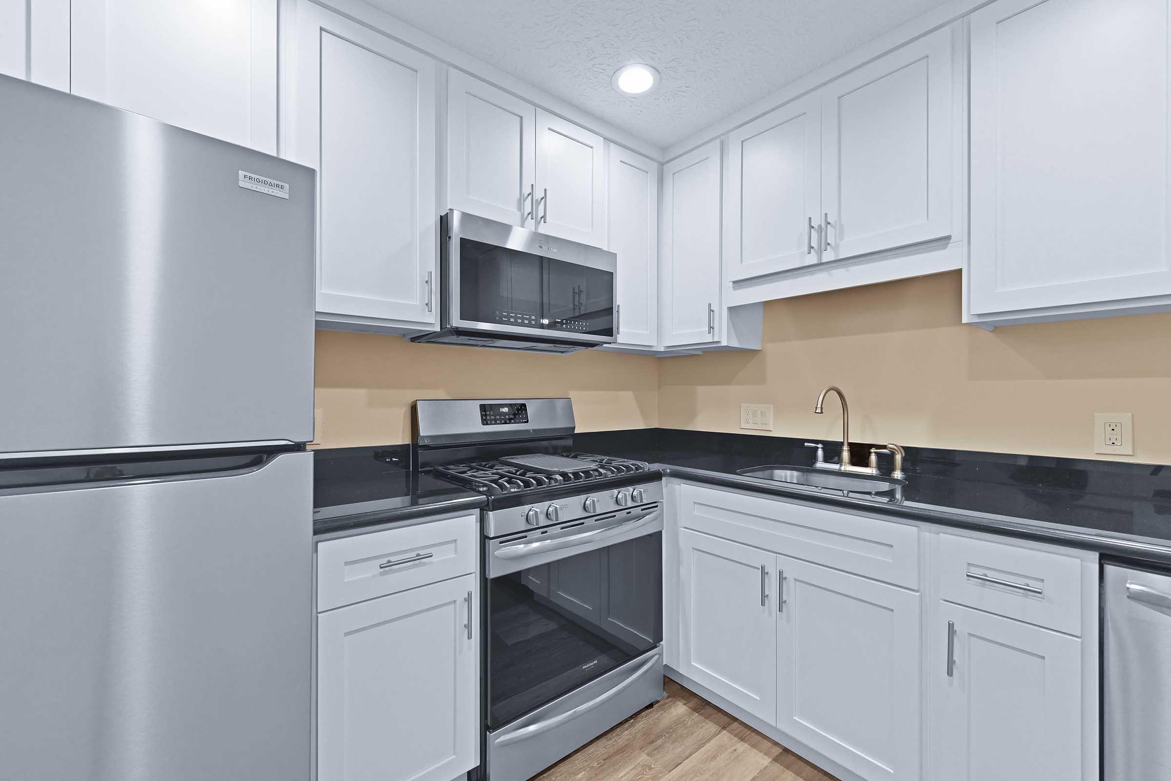 Modern kitchen featuring white cabinets, stainless steel appliances including a refrigerator and gas stove, and a microwave. Black countertop contrasts with the cabinetry, and there's a small sink with a sleek faucet. The walls are painted a light beige, giving a bright and clean appearance.