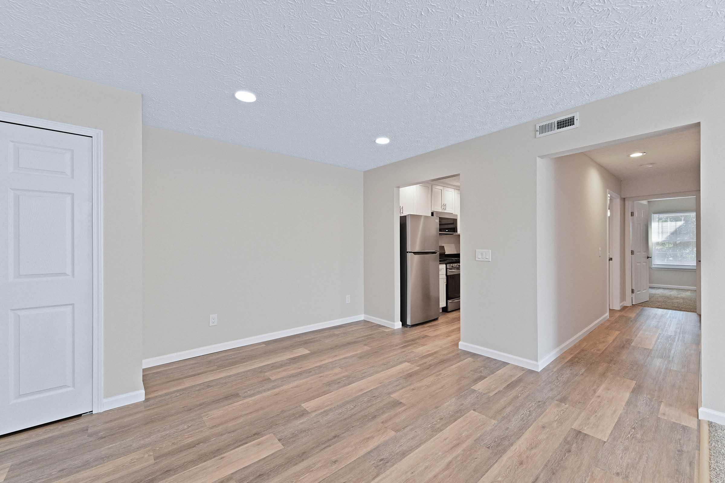 Spacious interior of a modern apartment featuring light-colored walls, hardwood-style flooring, and recessed lighting. To one side, a white door leads to another room, while the opposite side opens to a kitchen area with stainless steel appliances. Natural light fills the space, creating a bright ambiance.