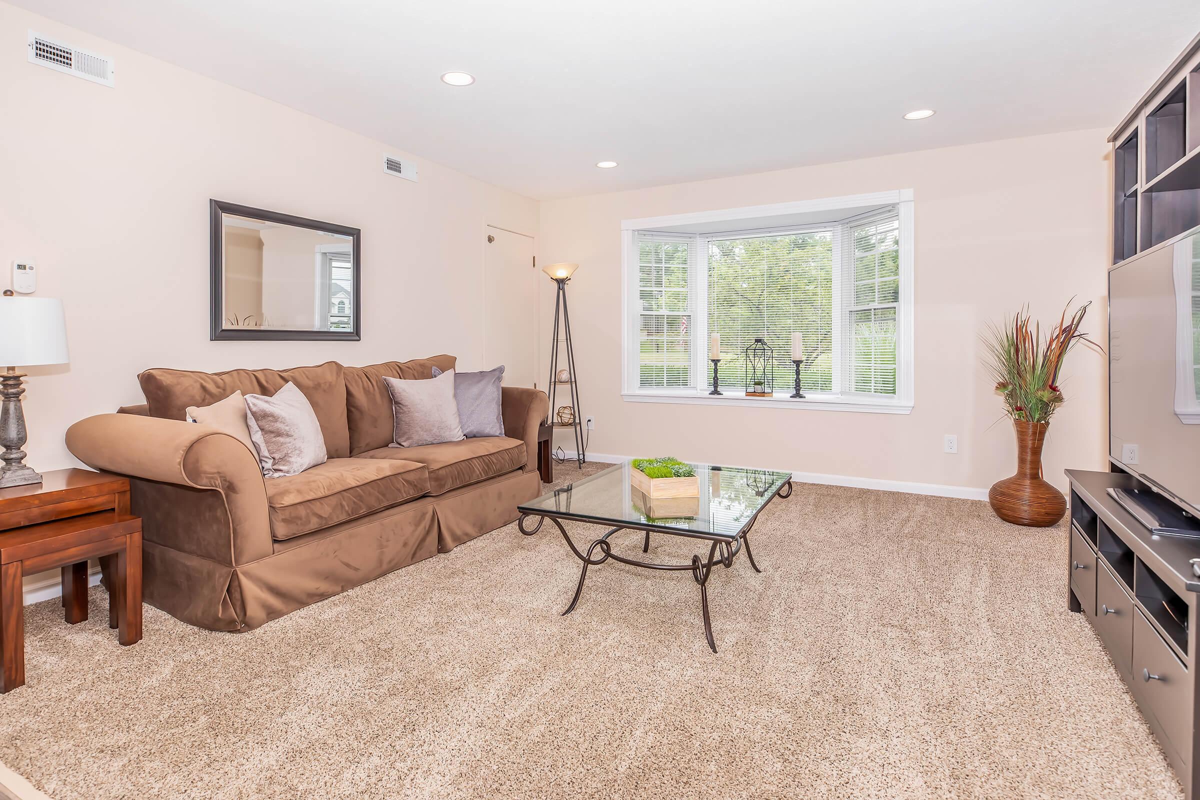 A cozy living room featuring a brown sofa with decorative pillows, a glass coffee table, and a floor lamp. The room has neutral wall colors, natural light streaming through a large window, and potted plants, creating a warm and inviting atmosphere.
