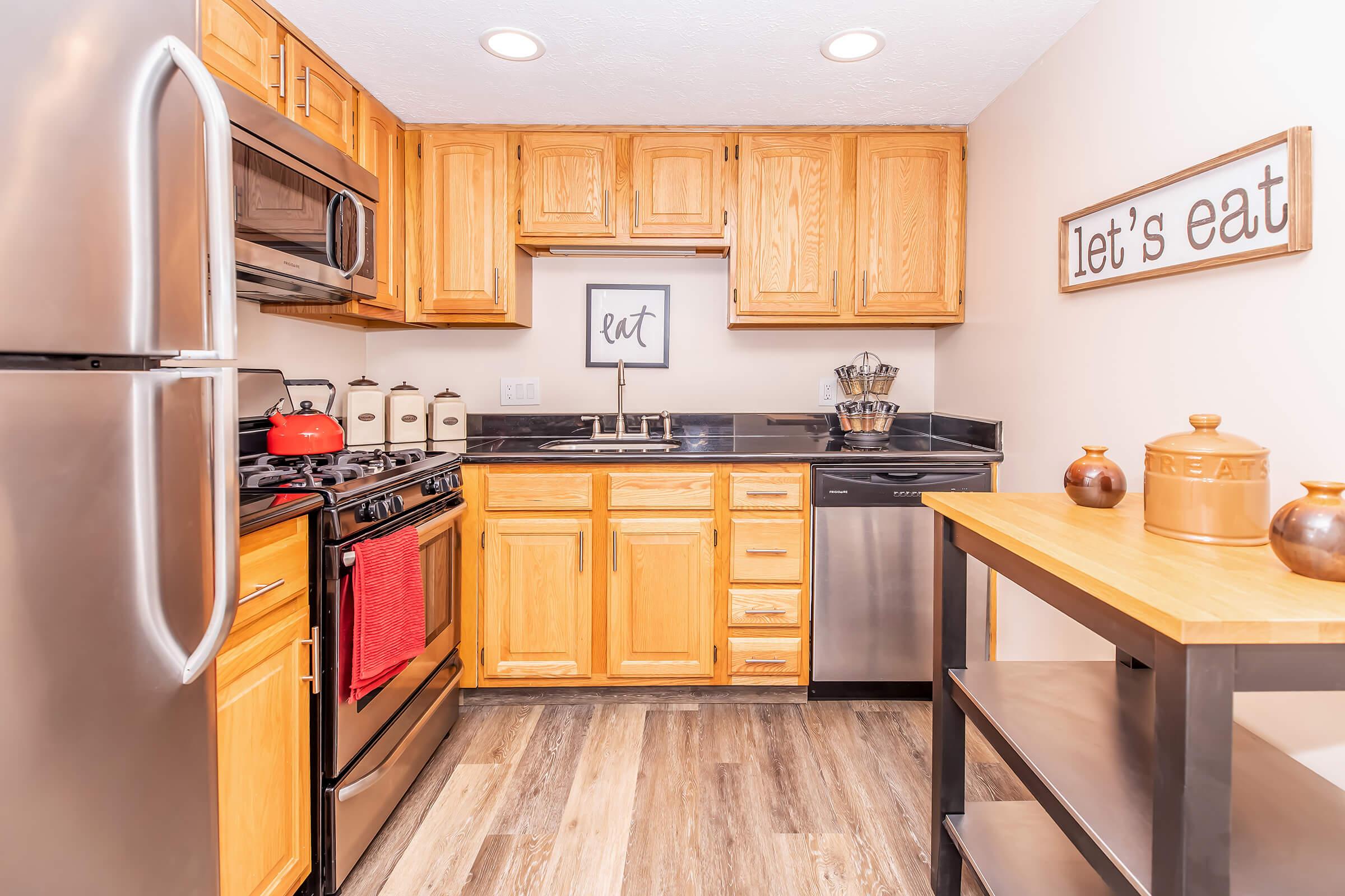 Modern kitchen featuring wooden cabinets, stainless steel appliances, and a black countertop. A red kettle sits on the stove, and decorative containers are arranged on the counter. A sign reading "let's eat" enhances the cozy atmosphere. The floor has a wood-like finish, and a small table holds two decorative jars.